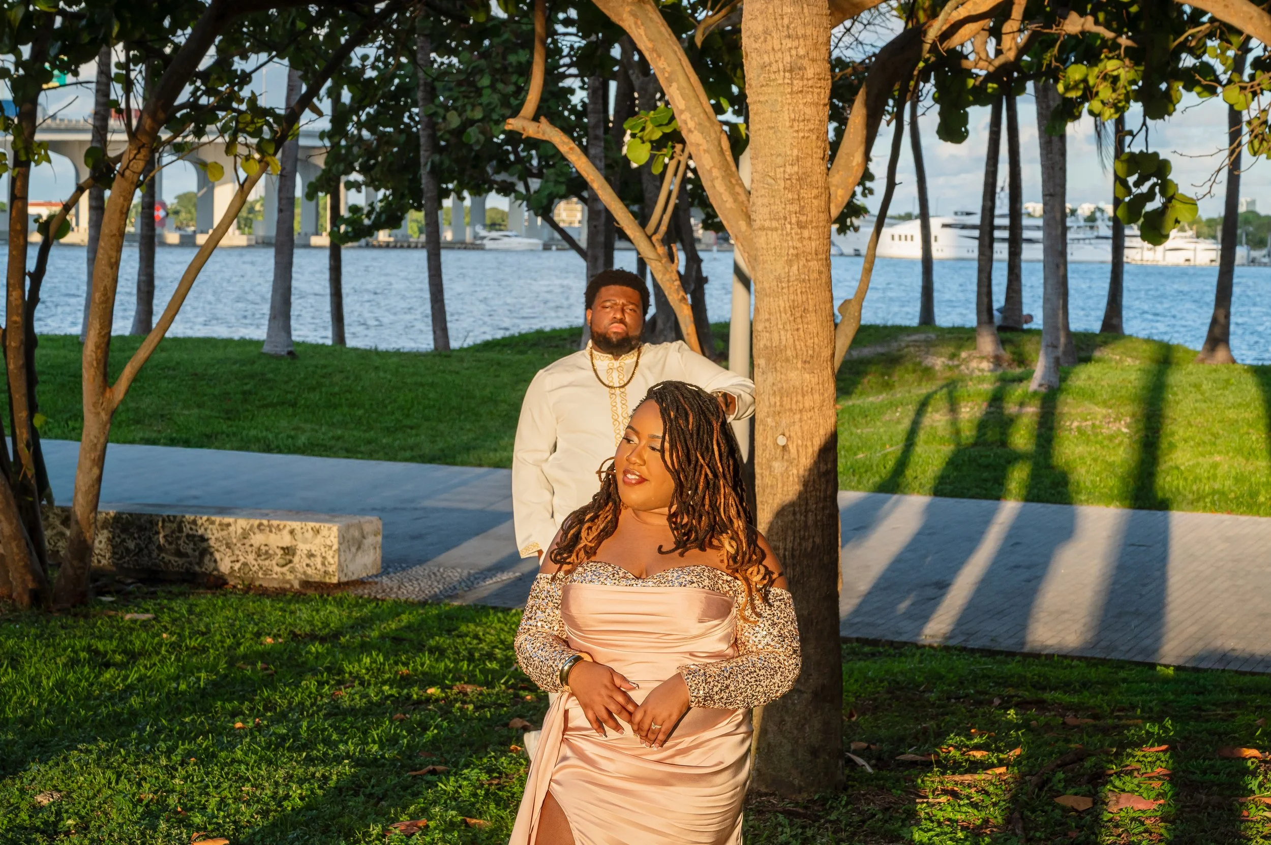 A woman with long, styled hair in a glamorous dress stands in front of a man in a white suit, both posing outdoors near a tree with water and yachts in the background during sunset.