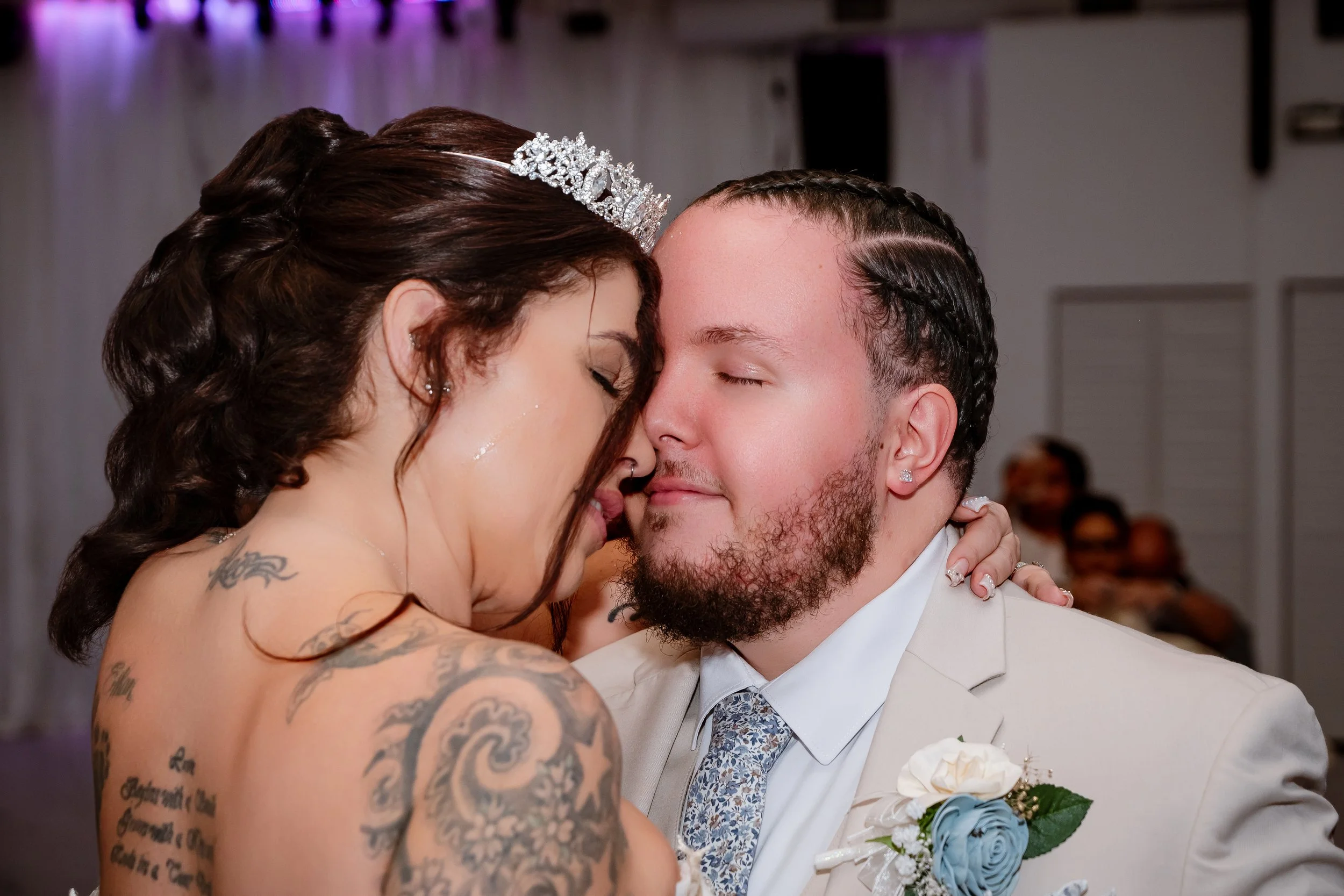 A bride and groom with their faces close together, eyes closed, during their wedding celebration. The bride has tattoos, jewelry, and a tiara, and the groom is dressed in a light-colored suit with a boutonniere.