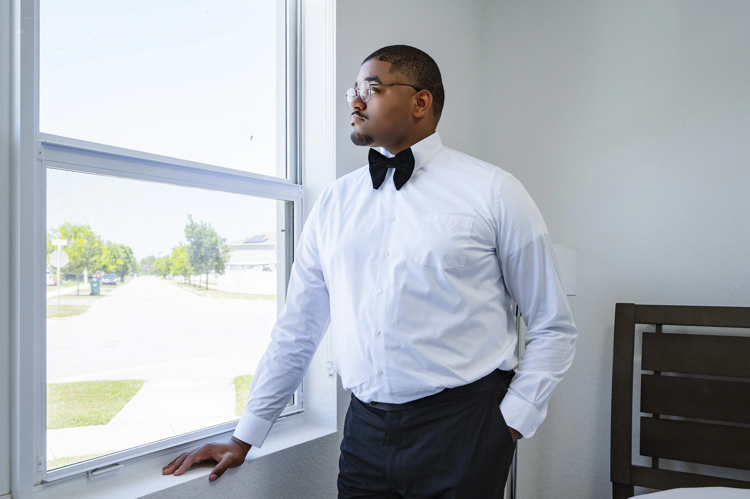 A man in a white shirt, black bow tie, and glasses standing by a window with natural light, in a simple room with a bed and a white wall.