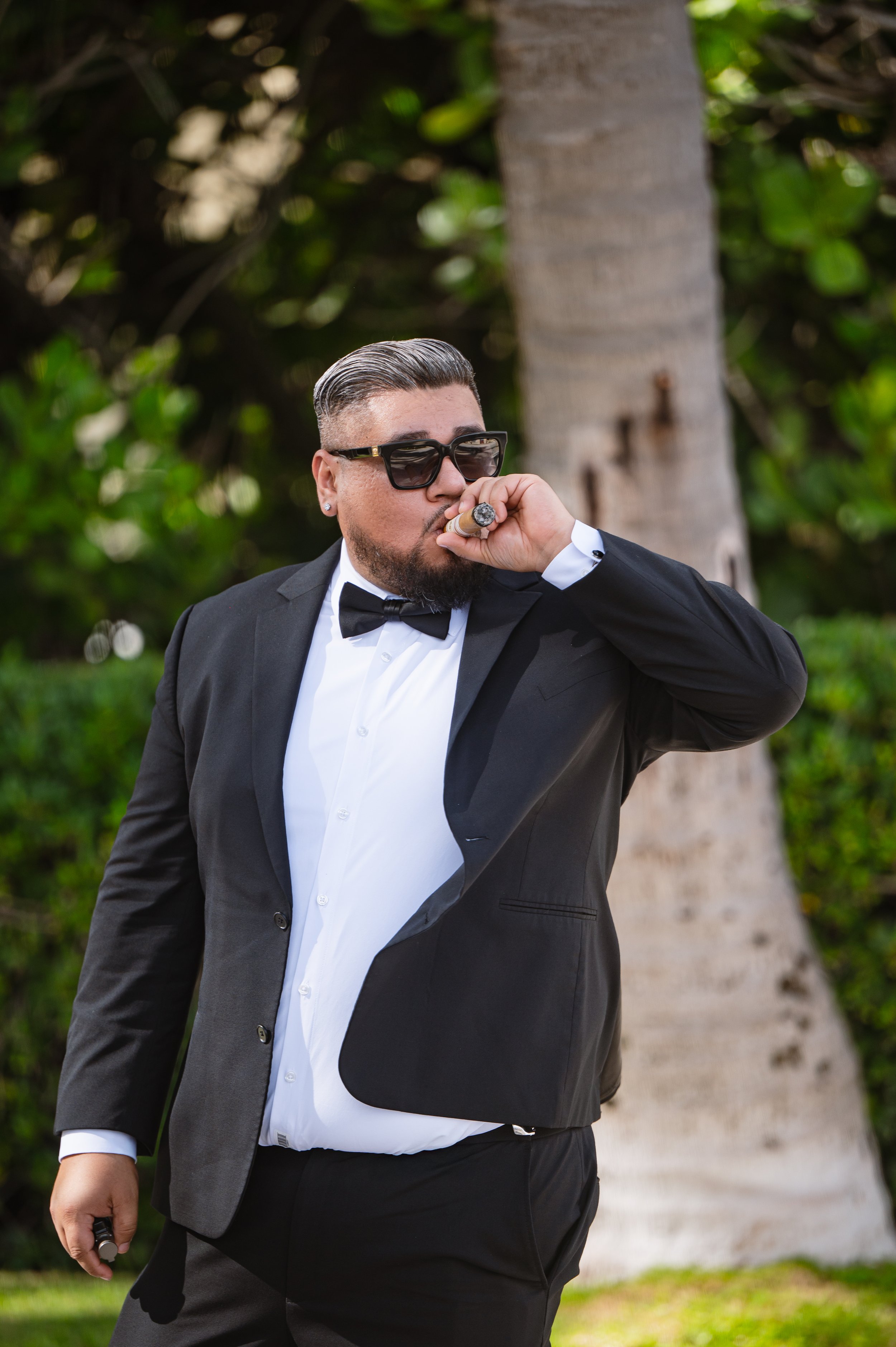 A man dressed in a black tuxedo, white shirt, and bowtie, wearing sunglasses, is smoking a cigar outdoors near a tree and greenery.