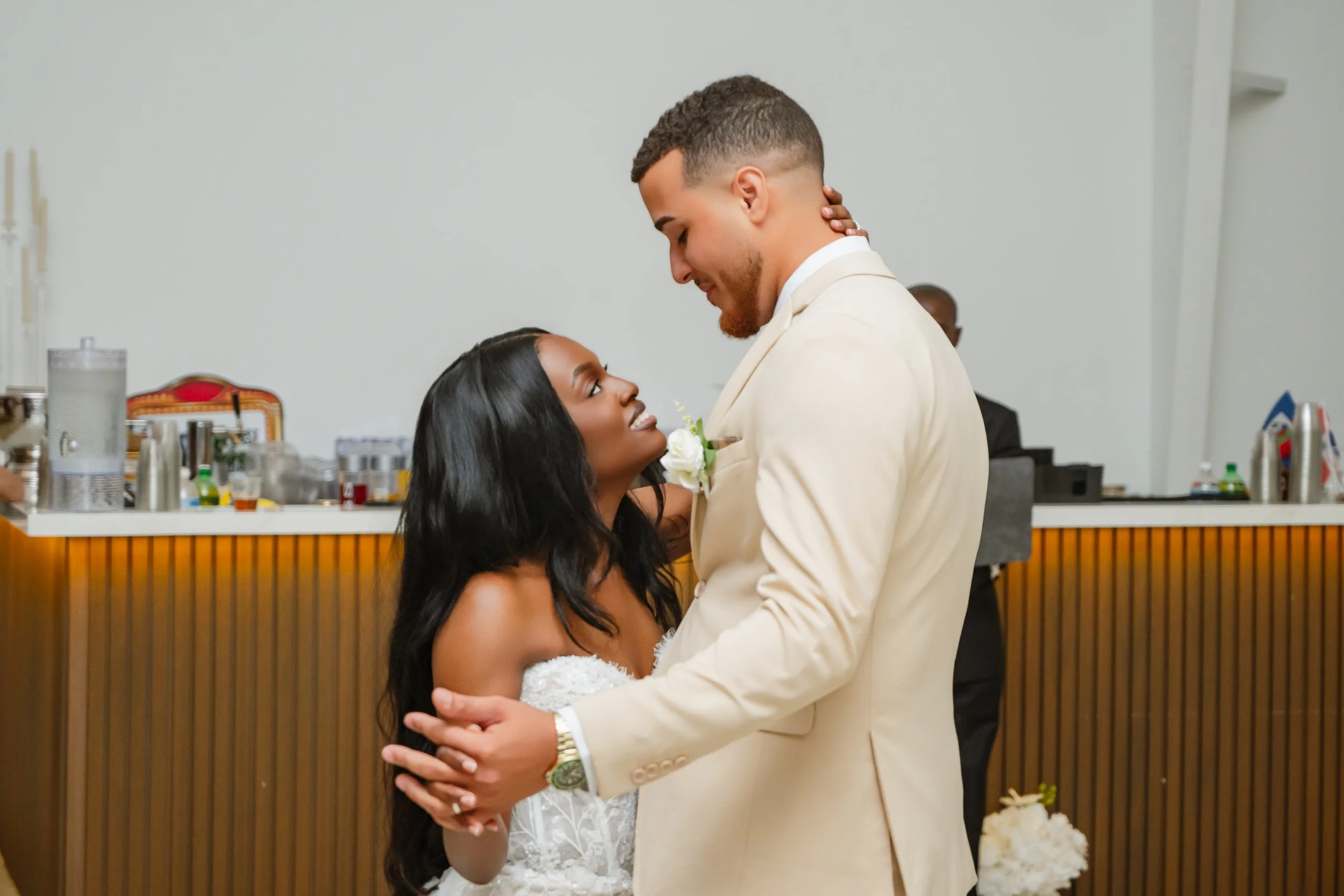 A bride and groom share a dance at their wedding reception, gazing into each other's eyes, with the bride wearing a white wedding dress and the groom in a cream-colored suit.