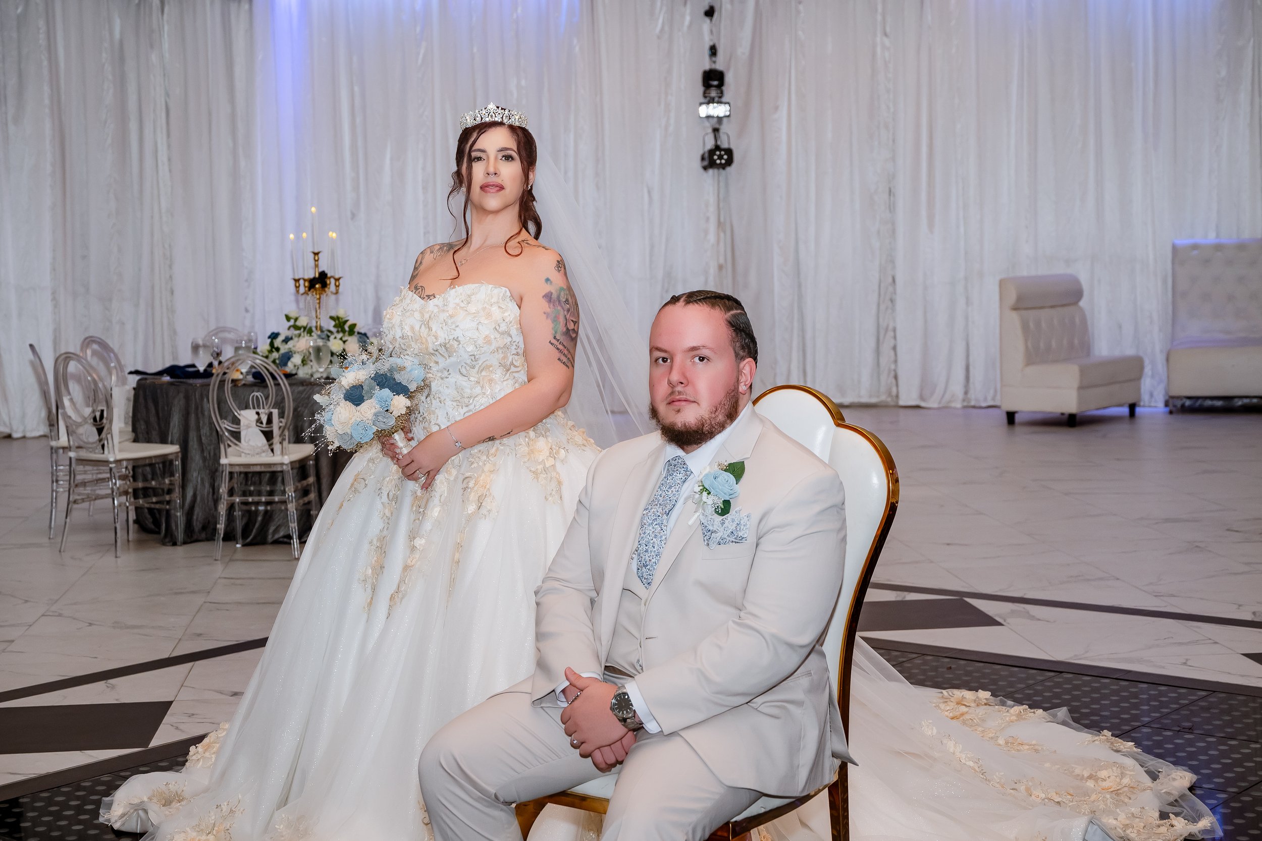 A bride and groom pose in wedding attire inside a decorated venue. The bride holds a bouquet of light blue and white flowers, wearing a tiara and veil, standing behind the seated groom in white suit with a boutonniere.