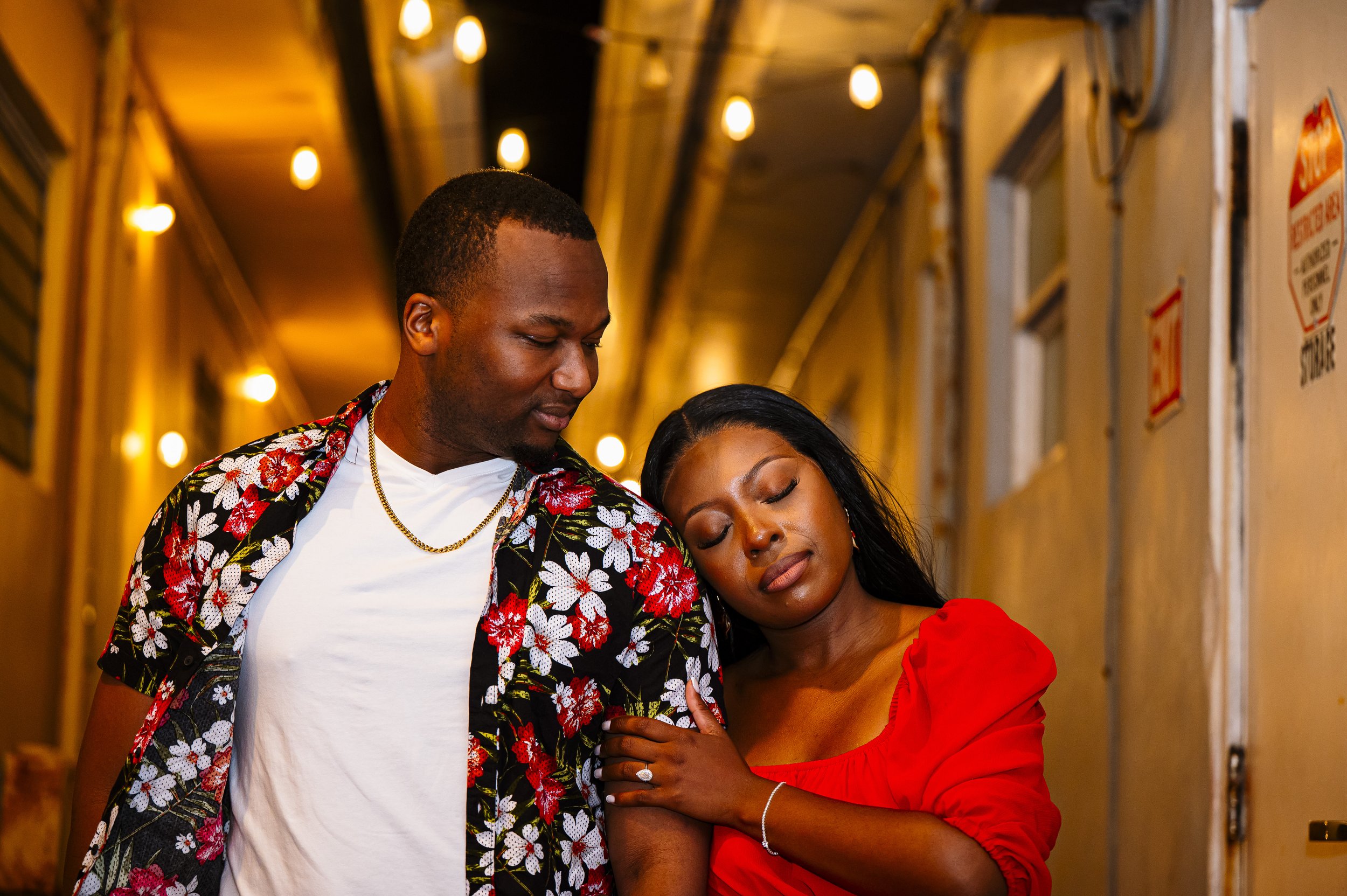 A man and woman standing close together in a warmly lit hallway, with the woman resting her head on the man's shoulder and holding his arm.