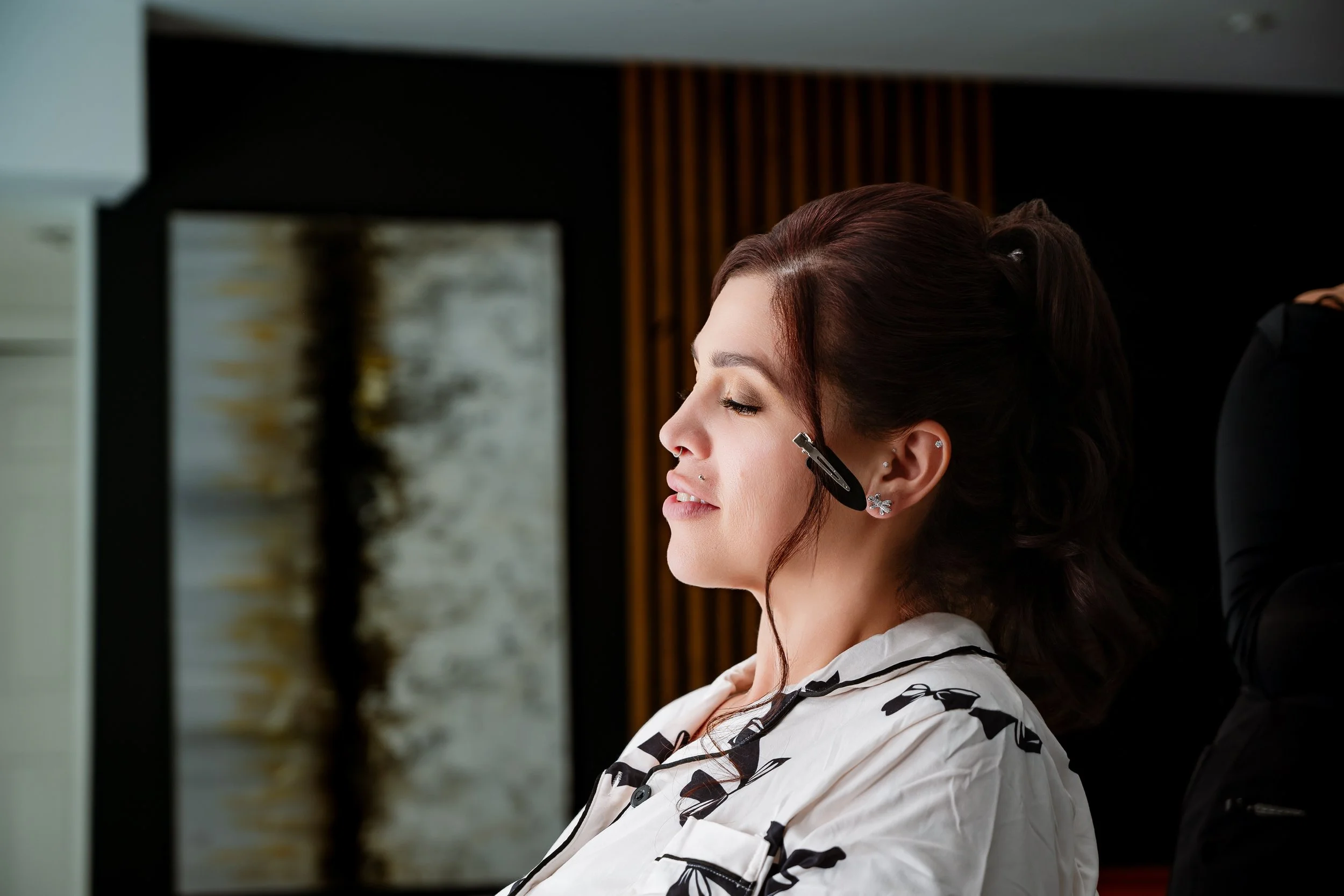A woman with closed eyes getting her hair styled, wearing a white robe with black bow patterns, and a black hair clip.