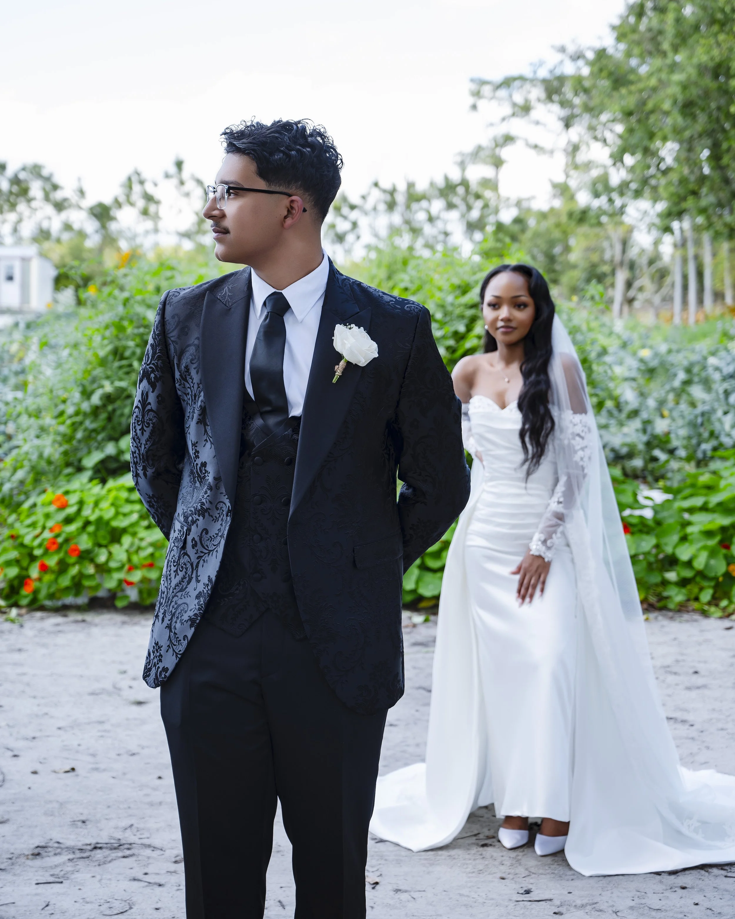 A groom in a black suit and tie with a white boutonniere stands with his back to his bride in a white wedding dress, on a sandy beach with green foliage in the background.