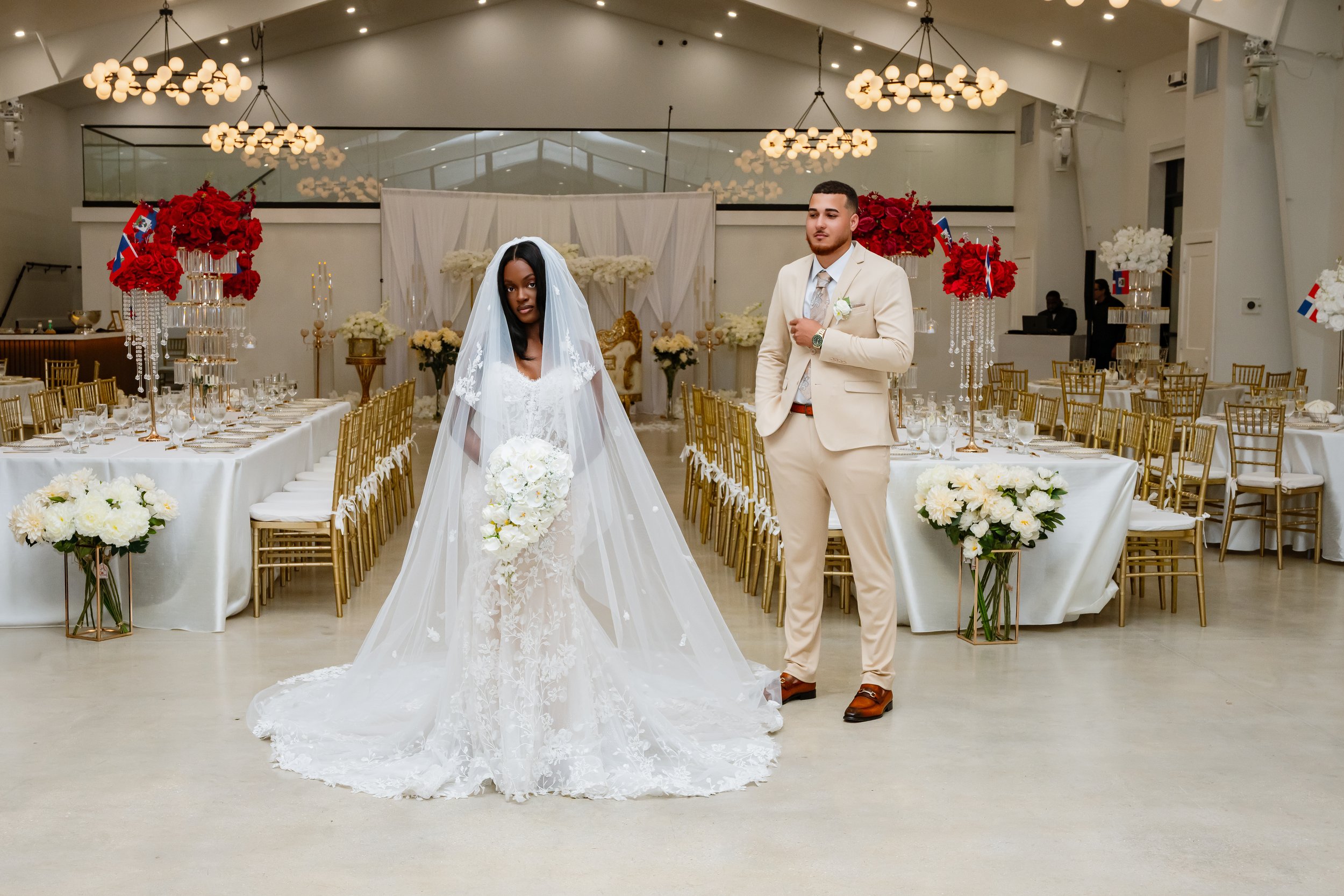 A bride in a white wedding gown and veil holding a bouquet of white flowers, standing next to a groom in a cream suit with a boutonnière, in a decorated wedding reception hall with tables, gold chairs, and floral centerpieces.