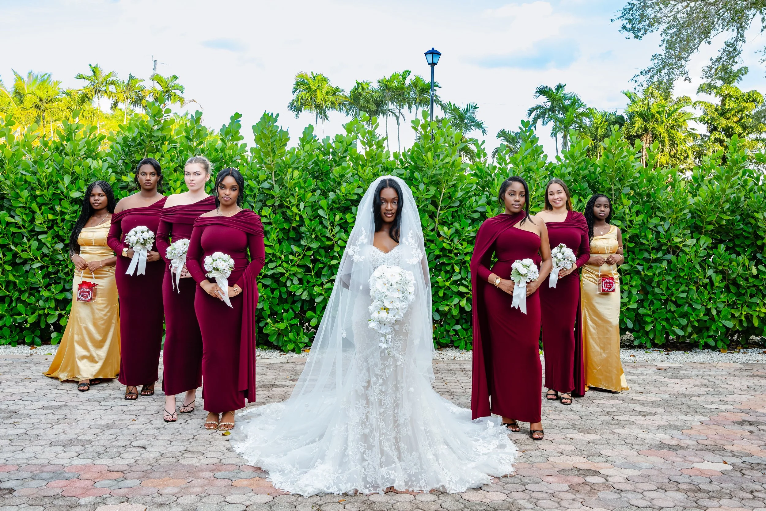 A bride in a white wedding dress and veil stands in the center holding a bouquet of white flowers, surrounded by six bridesmaids in maroon and gold dresses holding matching bouquets, outdoors with green bushes, palm trees, and a lamppost in the backg