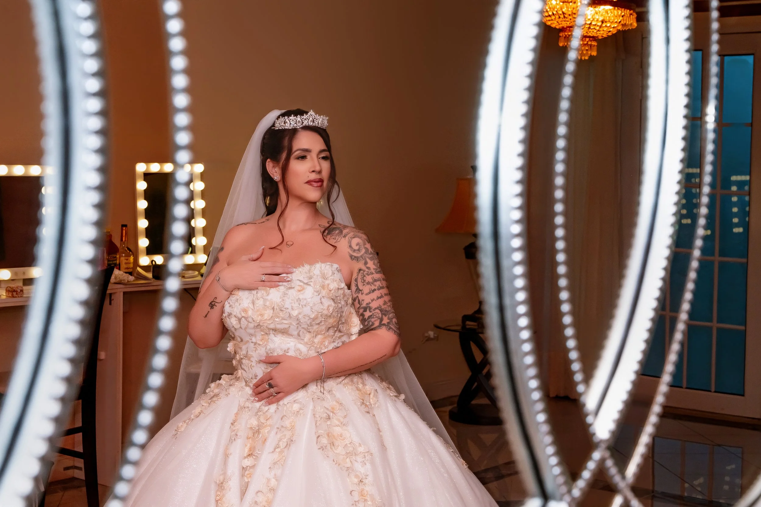Bride in a wedding dress with tattoos, wearing a crown and veil, posing with her hand on her chest in a warmly lit room, seen through decorative elements in the foreground.