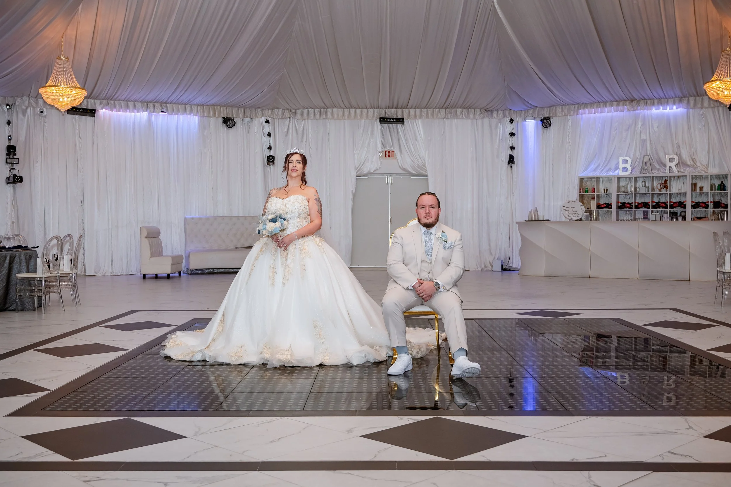 Bride in a white wedding gown with gold accents holding a bouquet standing next to groom in a white suit who is seated on a chair in a decorated wedding reception hall with white curtains and chandeliers.