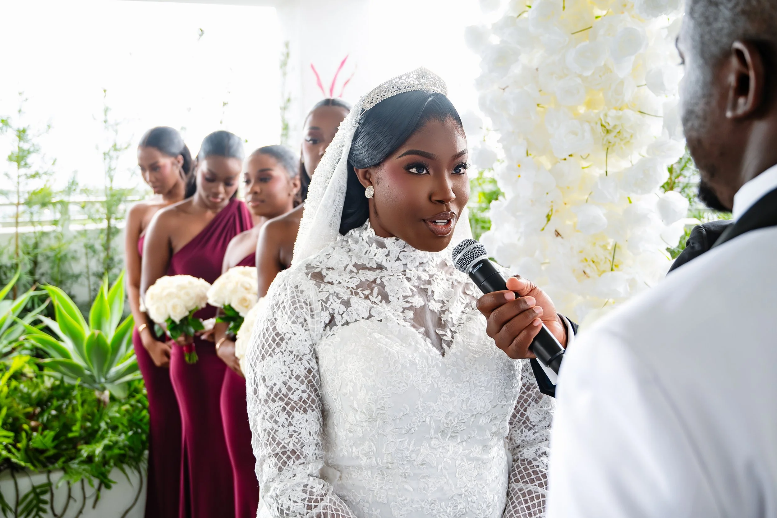 A bride in white lace dress and tiara holding a bouquet of white roses during her wedding ceremony, speaking into a microphone; lined with bridesmaids in burgundy dresses in the background.