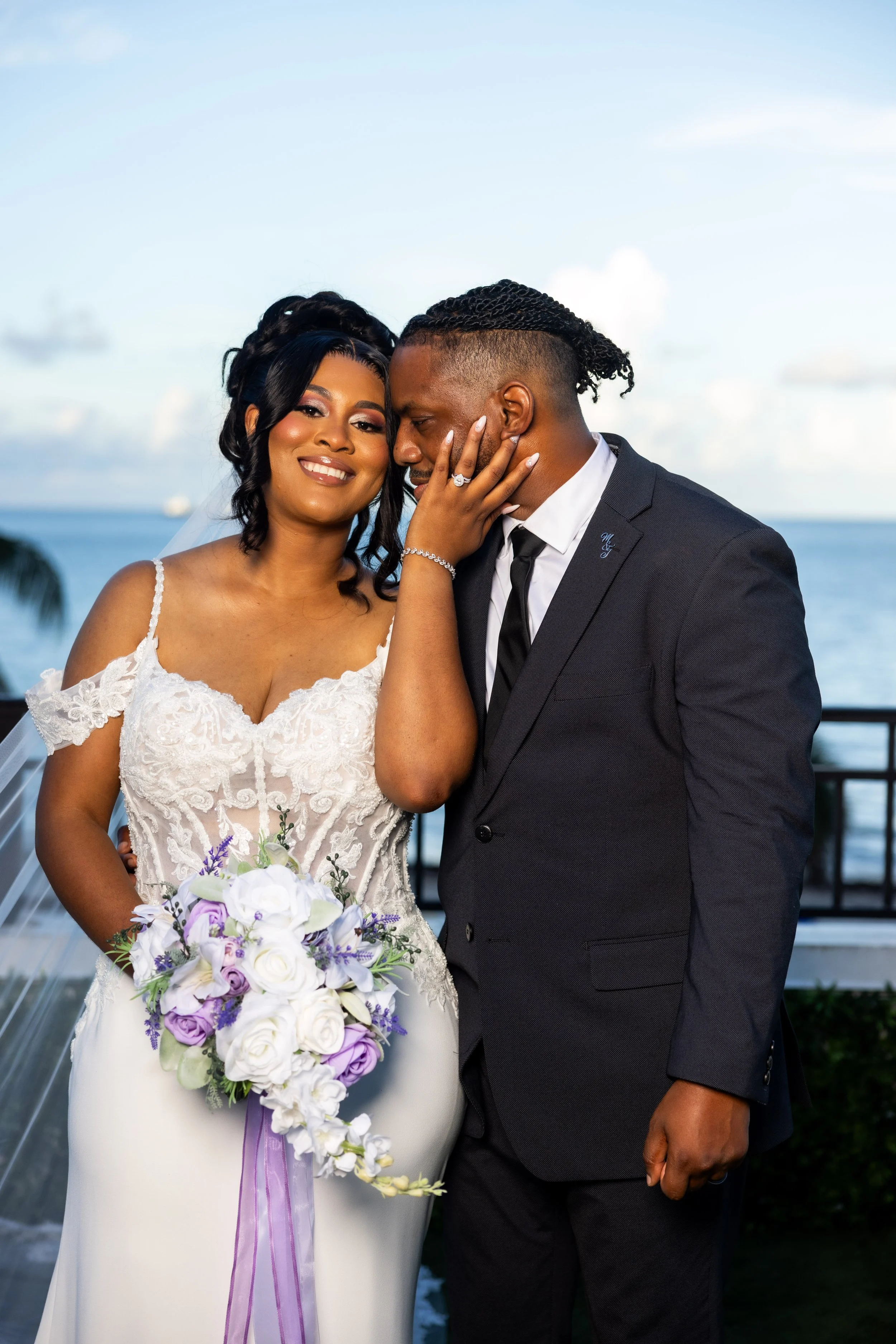 A bride and groom embrace during their outdoor wedding by the water, with blue sky and clouds in the background. The bride wears a lace wedding dress and holds a bouquet of white and purple flowers, while the groom is in a dark suit and tie.