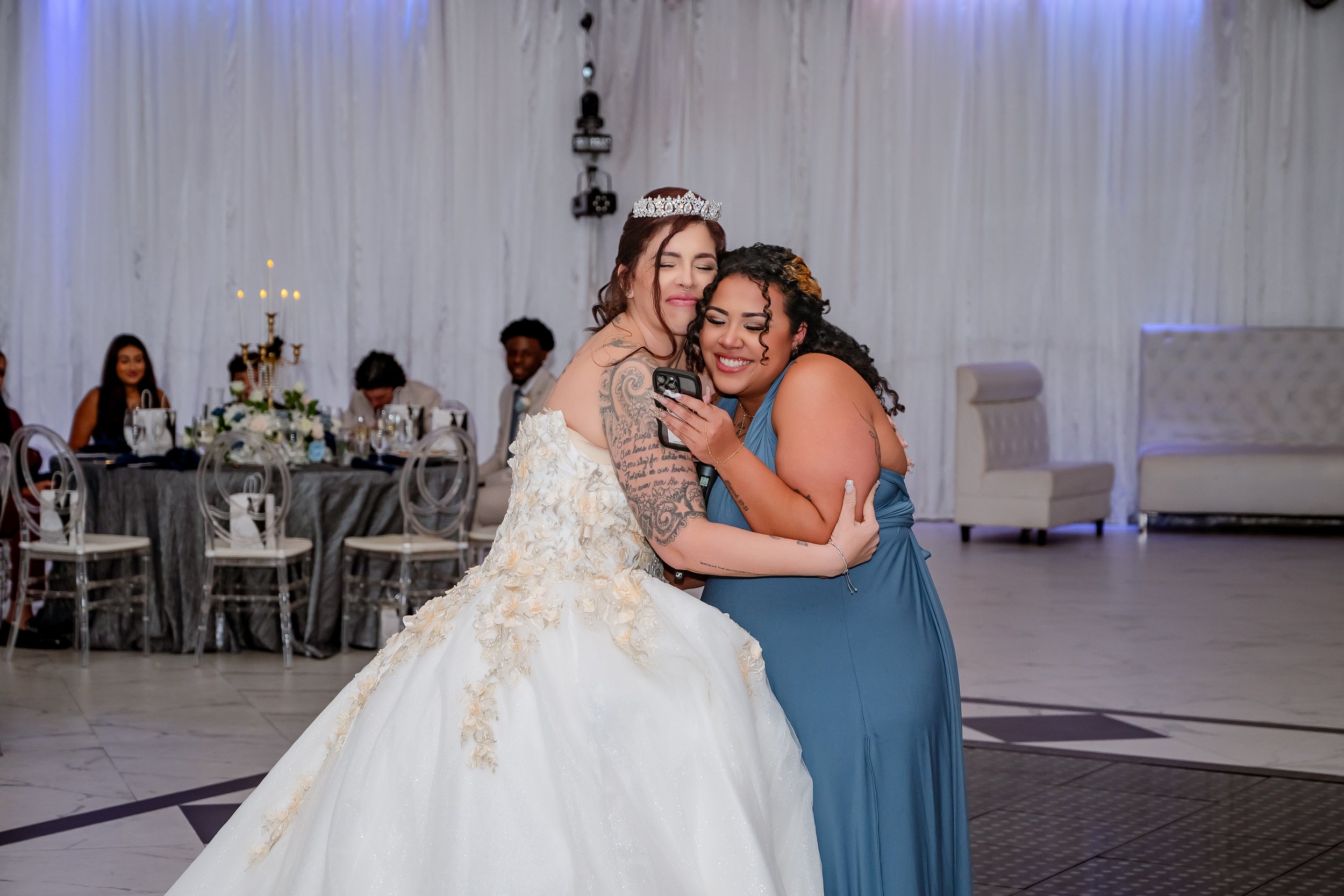 Two women hugging and smiling during a wedding reception; one woman is wearing a white wedding gown with a crown, and the other is in a blue dress, both happily looking at a cellphone.
