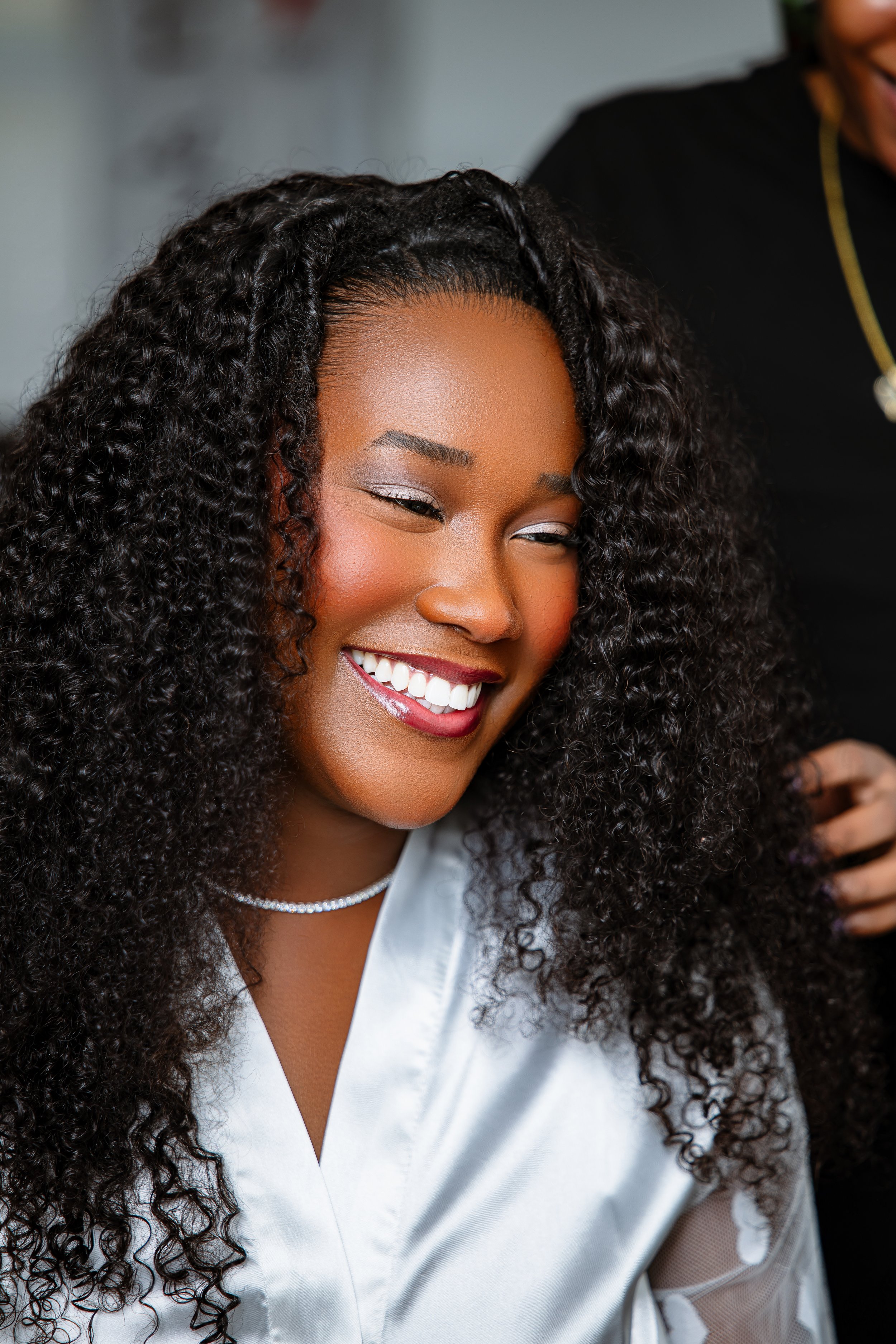 Close-up of a smiling African American woman with curly hair, wearing a white blouse and a pearl necklace, possibly getting ready or at a special event.