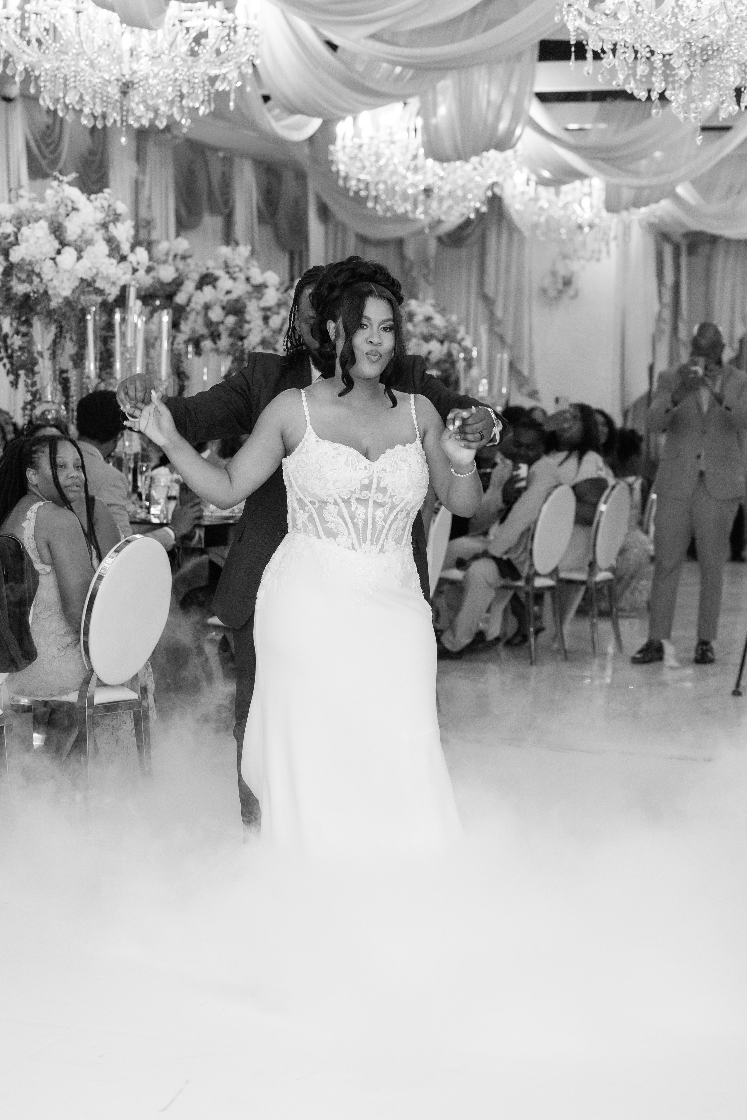A woman in a wedding dress dances with a man in a suit at a wedding reception, surrounded by guests seated at decorated tables with floral arrangements and chandeliers overhead.