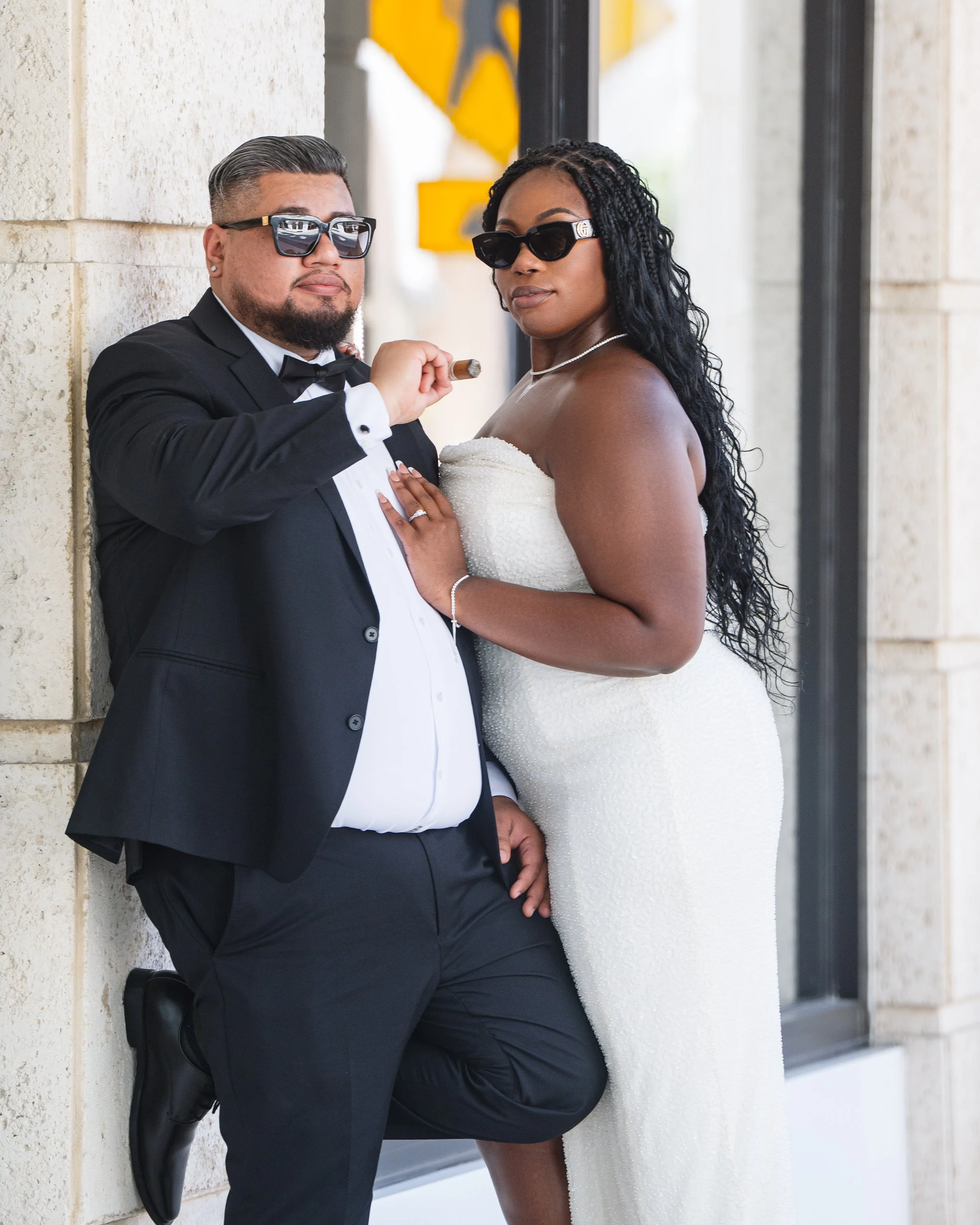 A couple dressed in formal attire, the man in a tuxedo with sunglasses holding a cigar and the woman in a strapless white dress with sunglasses, leaning against a wall outside a building.