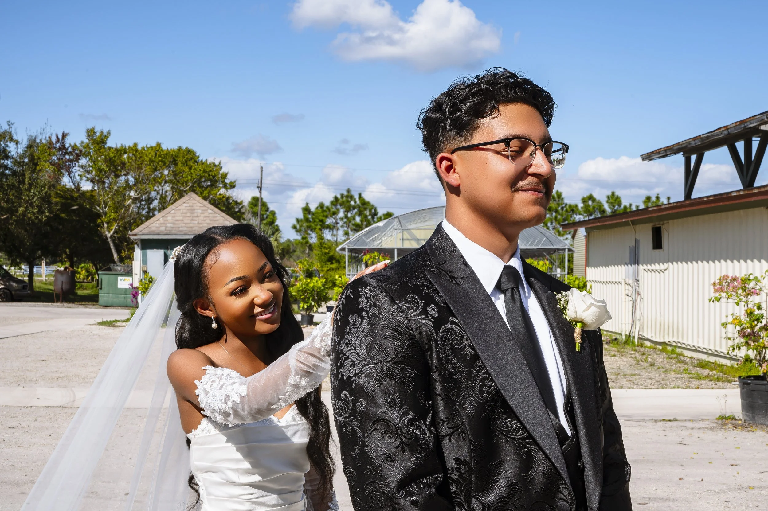 A bride and groom standing outdoors, with the bride smiling as she touches the groom's shoulder. The groom is dressed in a black suit with floral patterns, a white shirt, and a black tie, while the bride wears a white wedding gown with lace sleeves a
