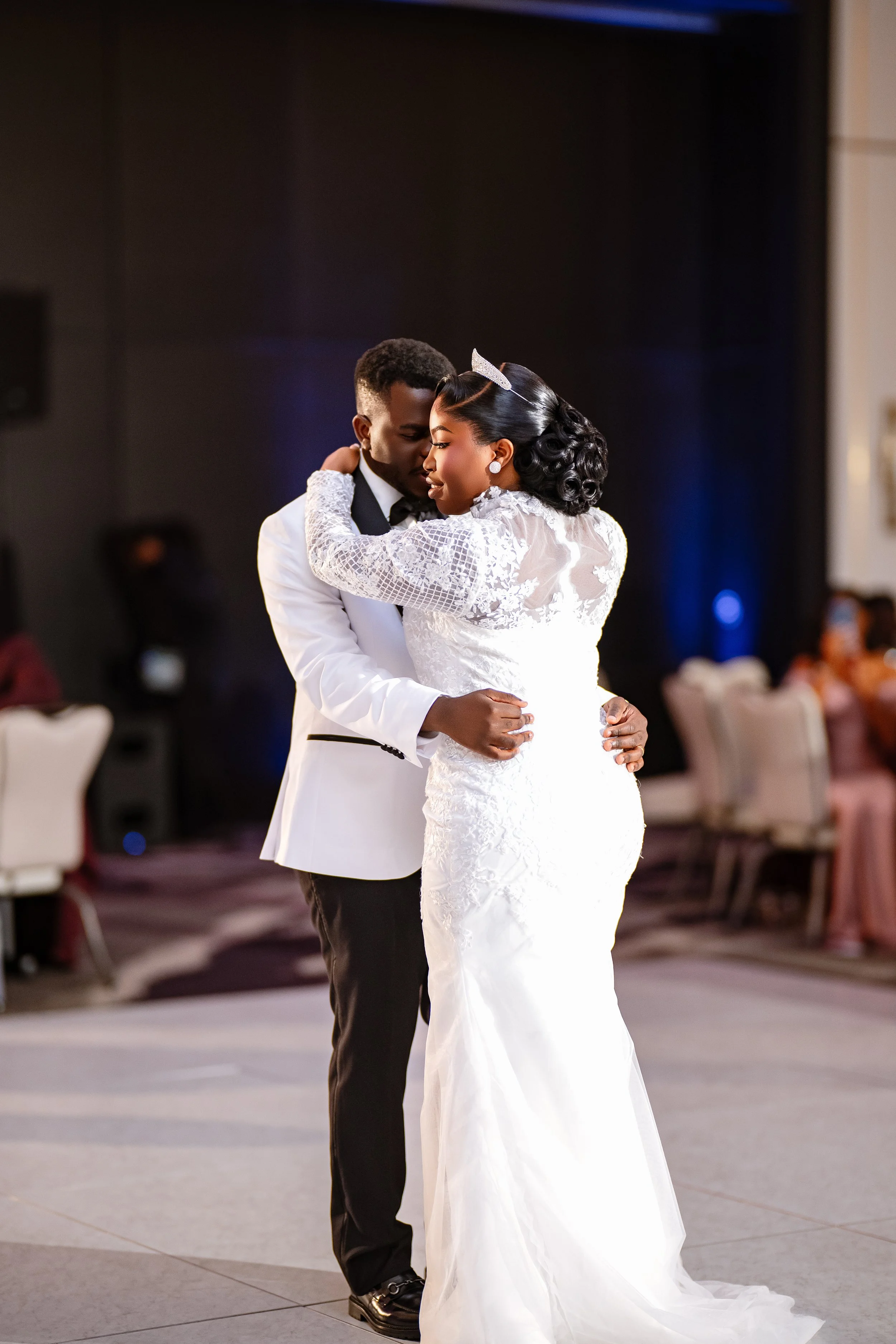 A couple dressed in wedding attire dancing closely during their wedding reception, with the bride in a white lace gown and the groom in a white tuxedo jacket with black pants.