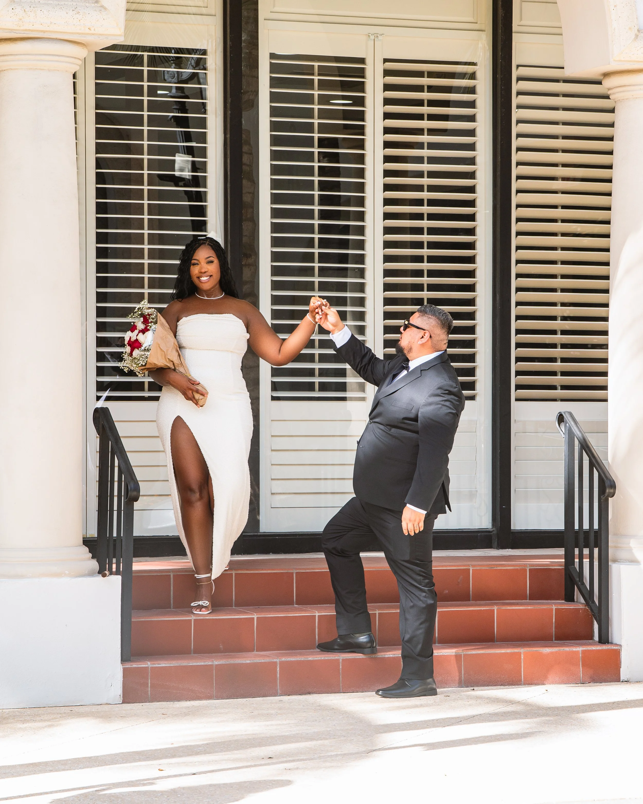 A smiling woman in a white dress with a high slit receives keys from a man in a black suit on a porch with stairs and white shutters.