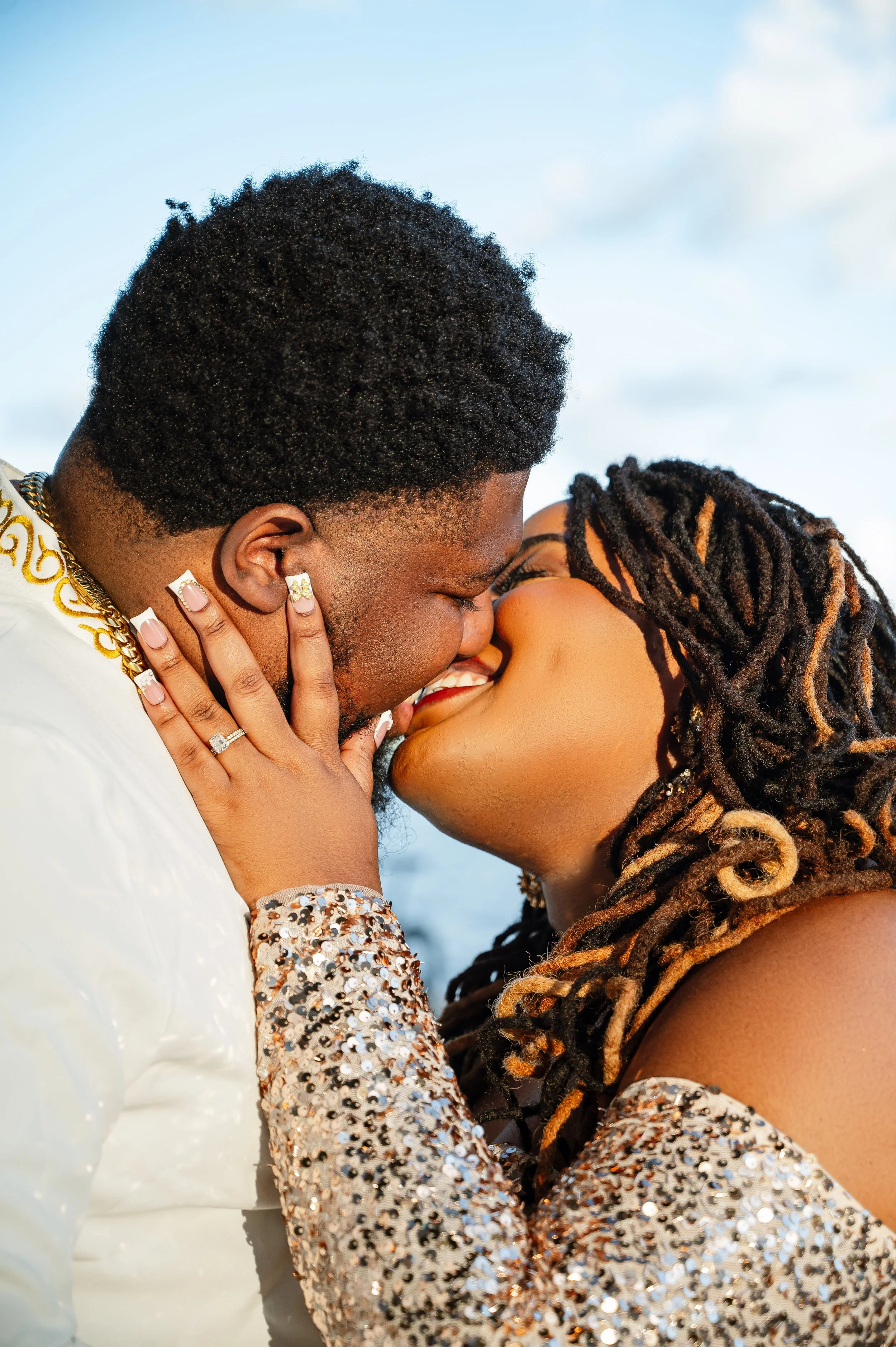 A couple is kissing outdoors, with both smiling and holding each other close. The man has a beard and wearing a white jacket with gold embellishments, while the woman has long braided hair and dressed in a shiny, sequined top.