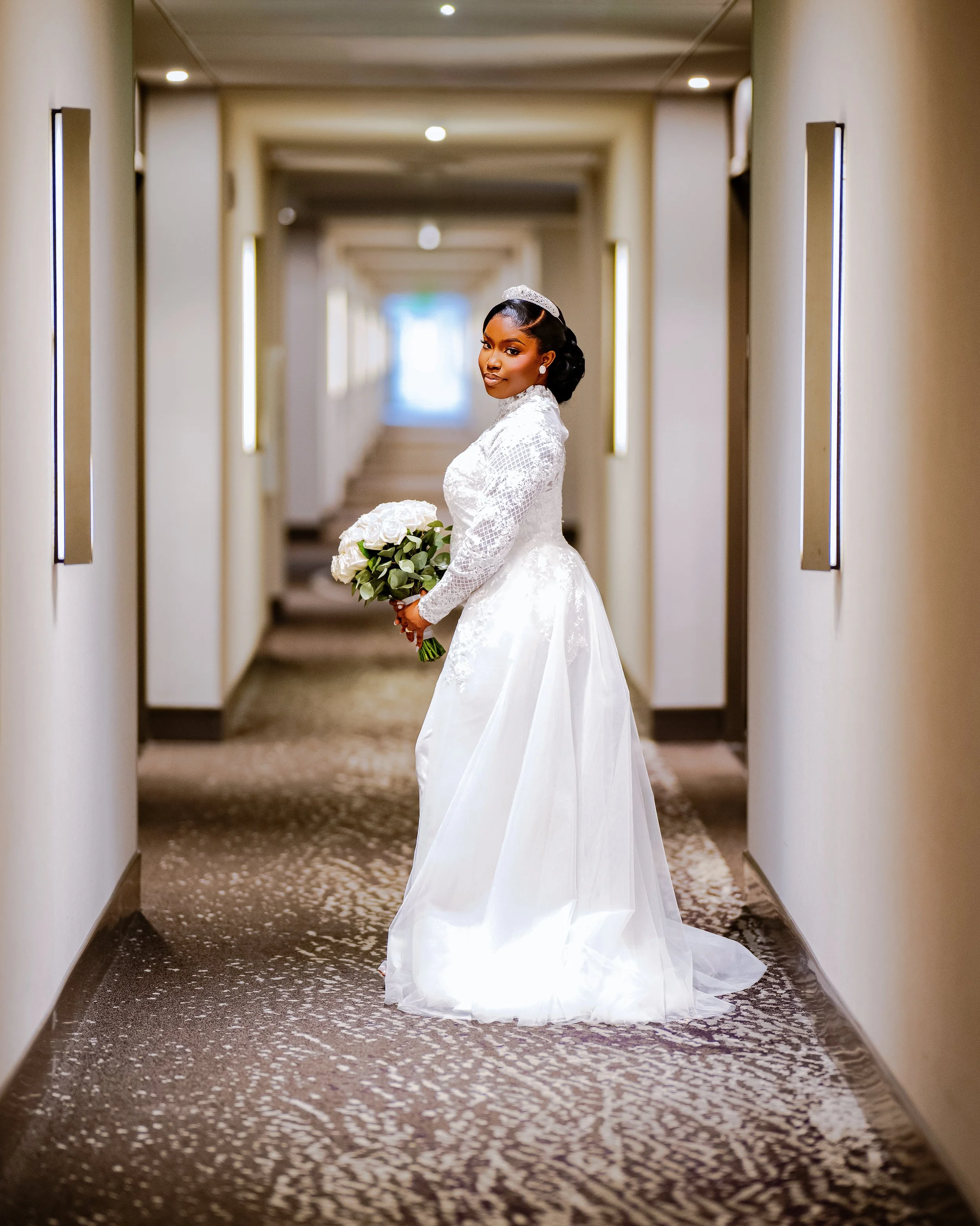 A bride in a white wedding dress holding a bouquet of white roses standing in a hallway with beige walls and illuminated rectangular wall lights.