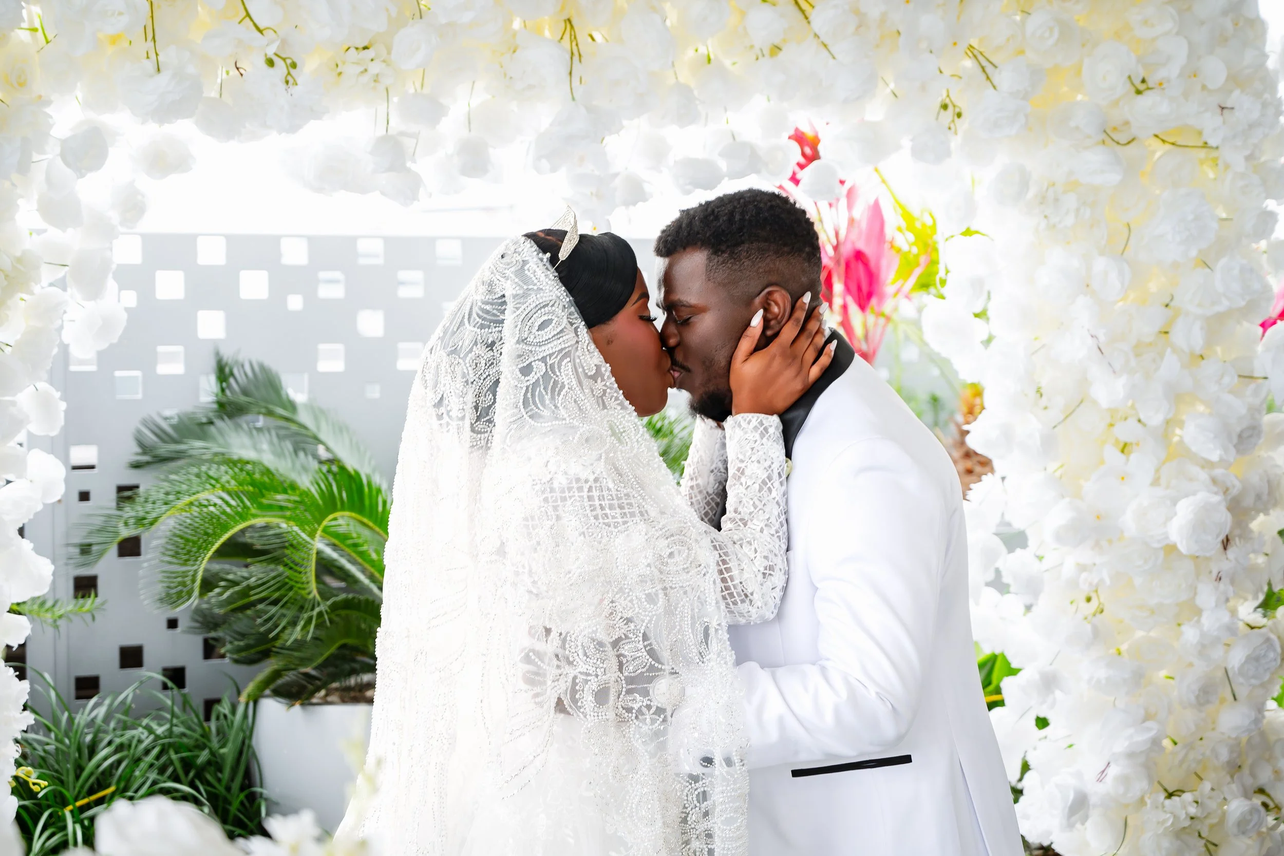 A bride and groom sharing a kiss at their wedding surrounded by white flowers and greenery.