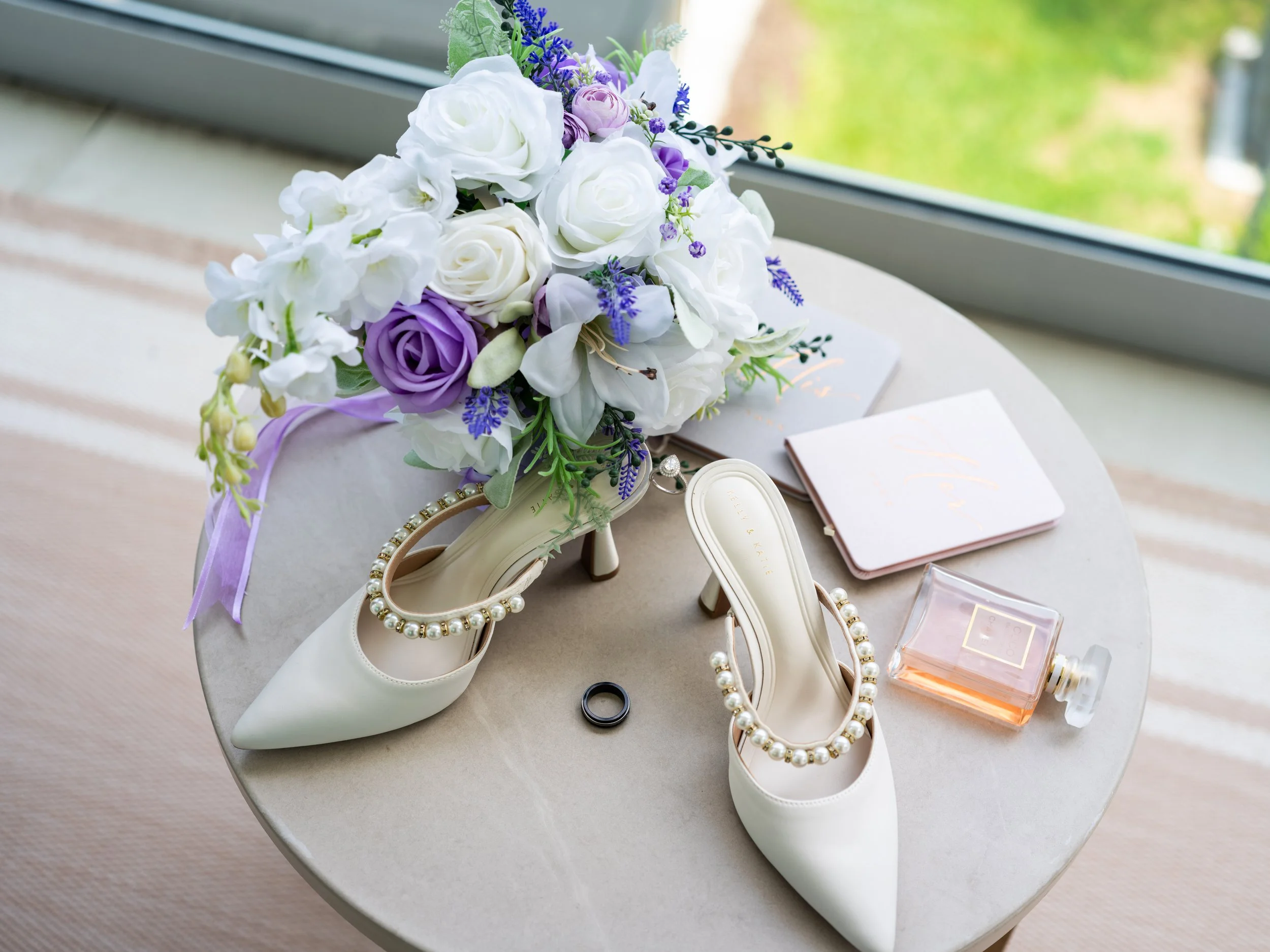 A bridal bouquet with white roses, purple roses, and lavender rests on a table. Surrounding it are white high heels with pearl embellishments, a pearl necklace, a wedding ring, a black ring, a jewelry box, a lipstick, and a book, with a window and gr