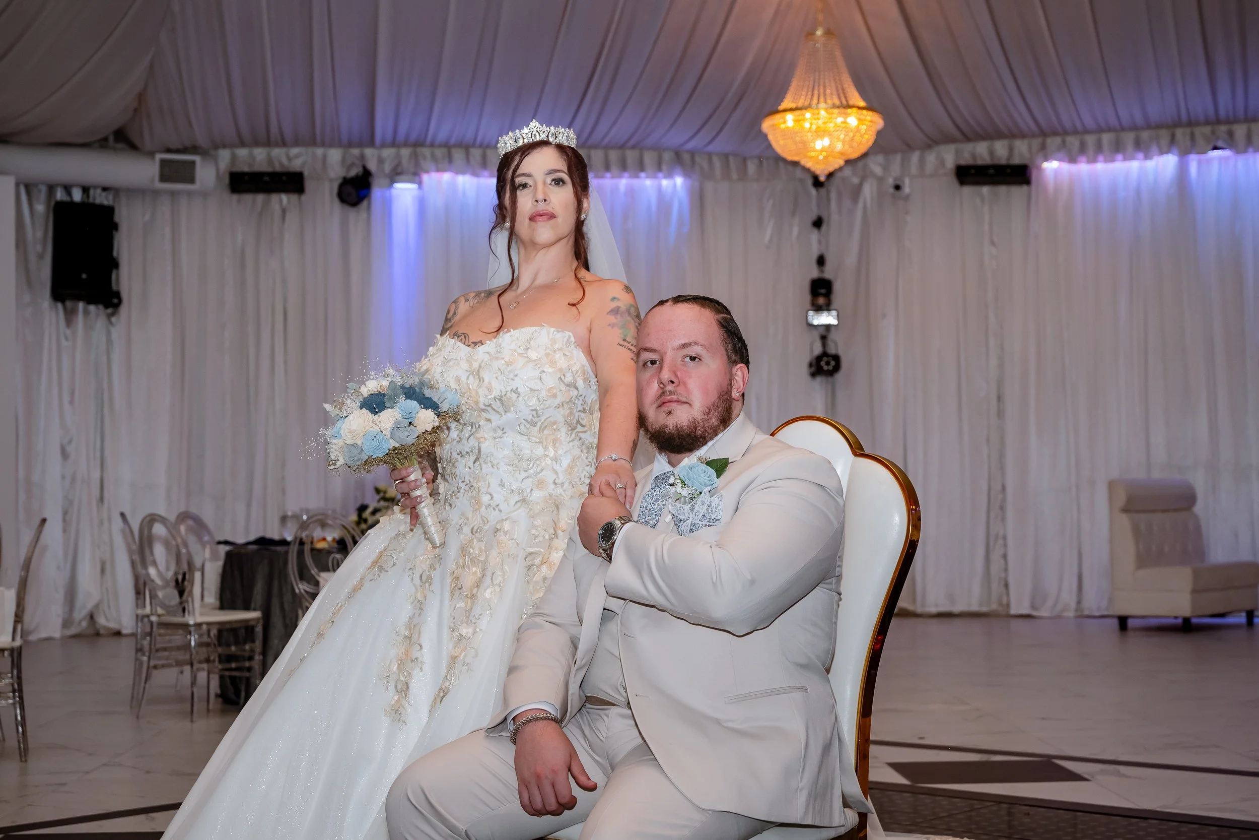 A bride and groom at their wedding reception. The bride is wearing a white strapless wedding gown, a tiara, and holding a bouquet of blue and white flowers. The groom is seated in a white tuxedo with a blue boutonniere, resting his arm on his knee. T
