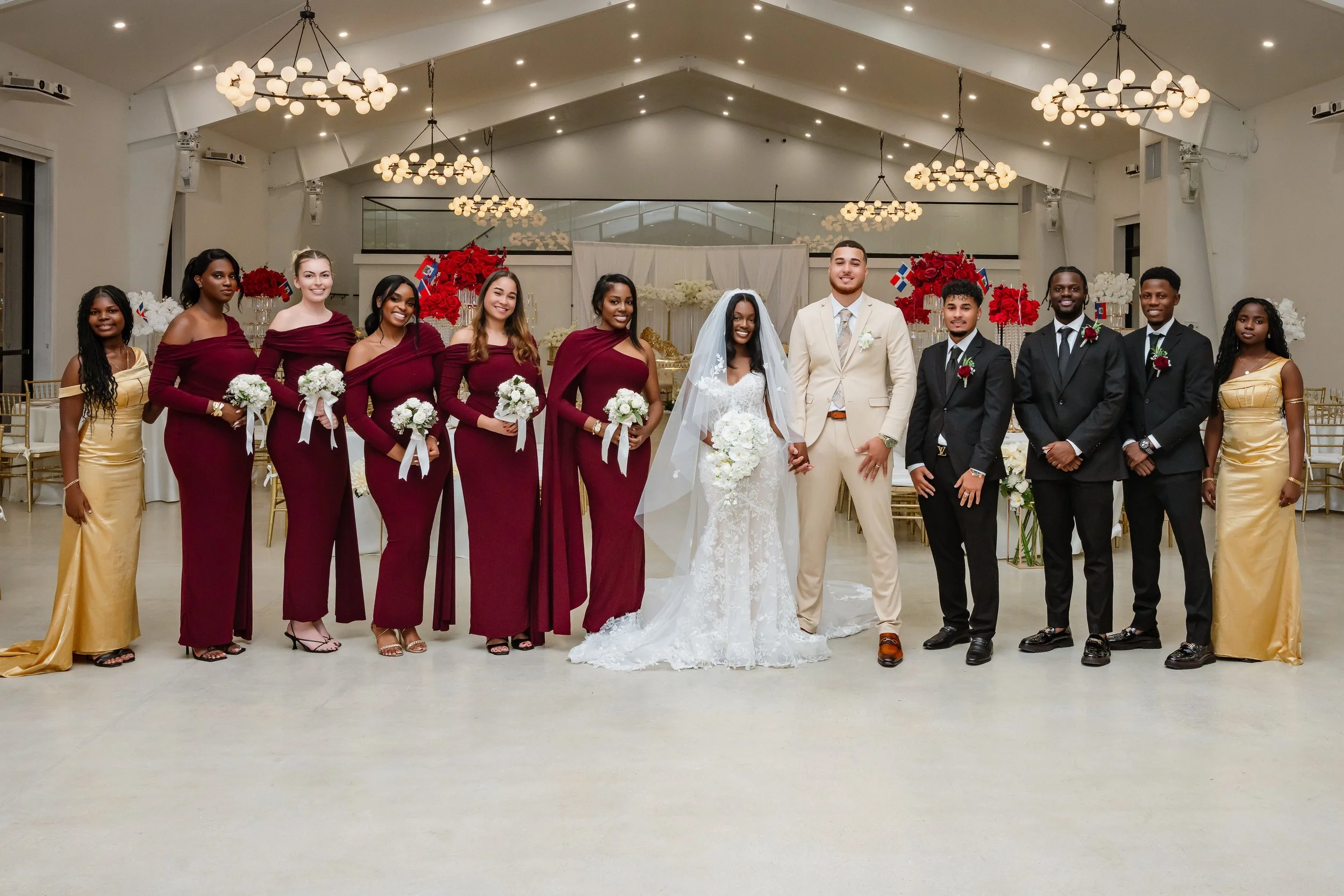 Wedding party standing inside a decorated event hall, including bride, groom, and their attendants dressed in formal attire. The bride is wearing a white lace wedding gown with veil, holding a bouquet. The groom is in a light beige suit with a white 