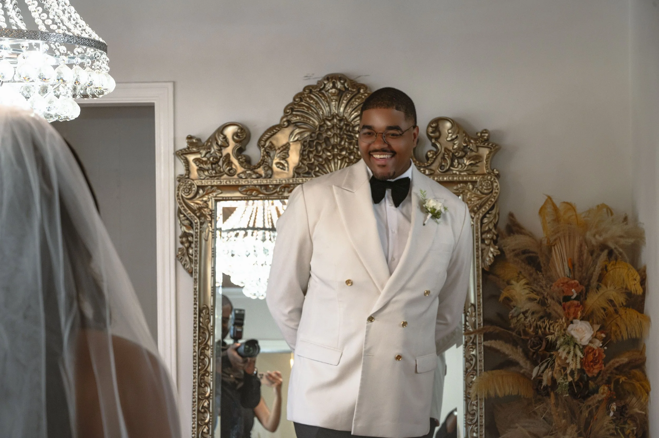 A man wearing a white tuxedo with a black bow tie and glasses, smiling, standing in front of an ornate gold mirror, with a flower boutonniere on his left lapel. A photographer is visible in the mirror's reflection.