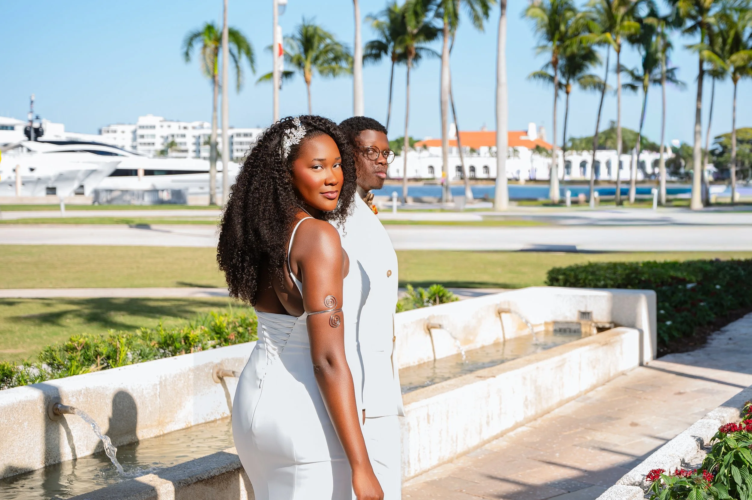 A couple dressed in white, standing beside a water fountain in a park with palm trees and white buildings in the background.