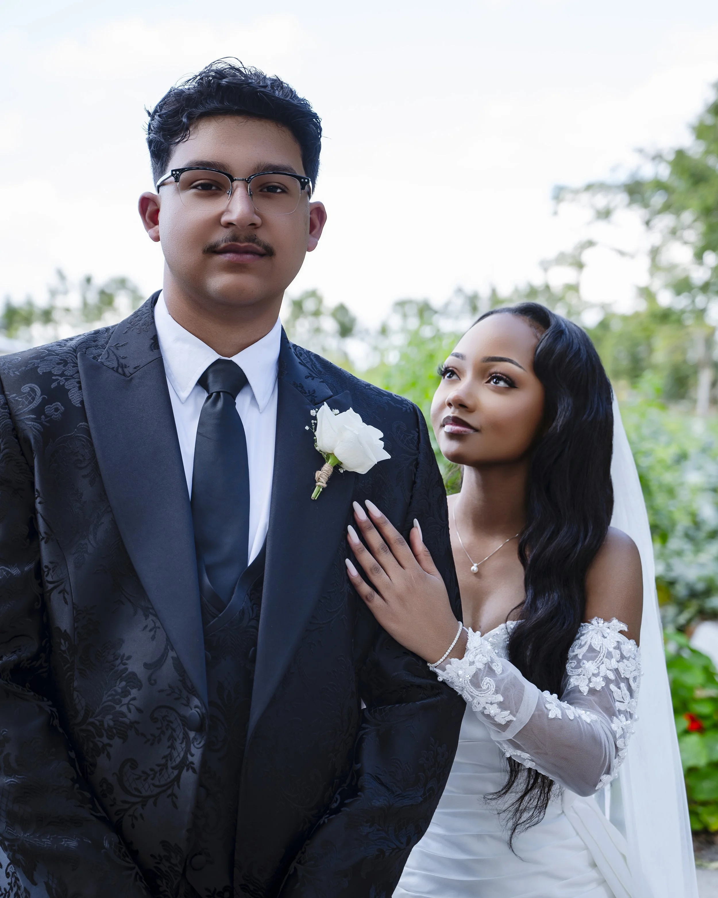 A bride and groom outdoors at wedding, groom wears suit with boutonniere, bride in wedding gown and veil, bride touching groom's arm, greenery in background.
