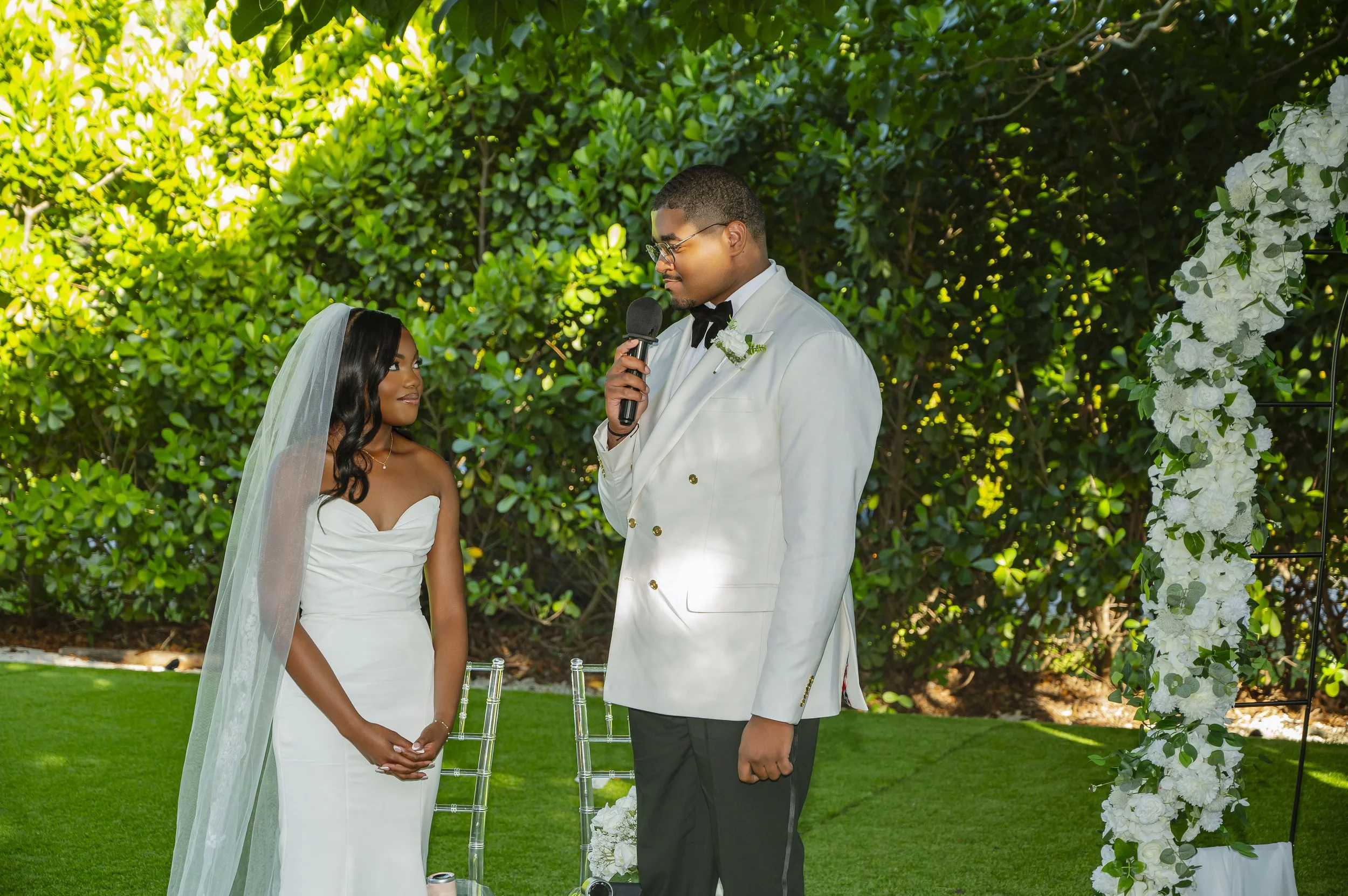 Bride and groom exchanging vows outdoors, with greenery in the background and a floral arch decoration to the right.