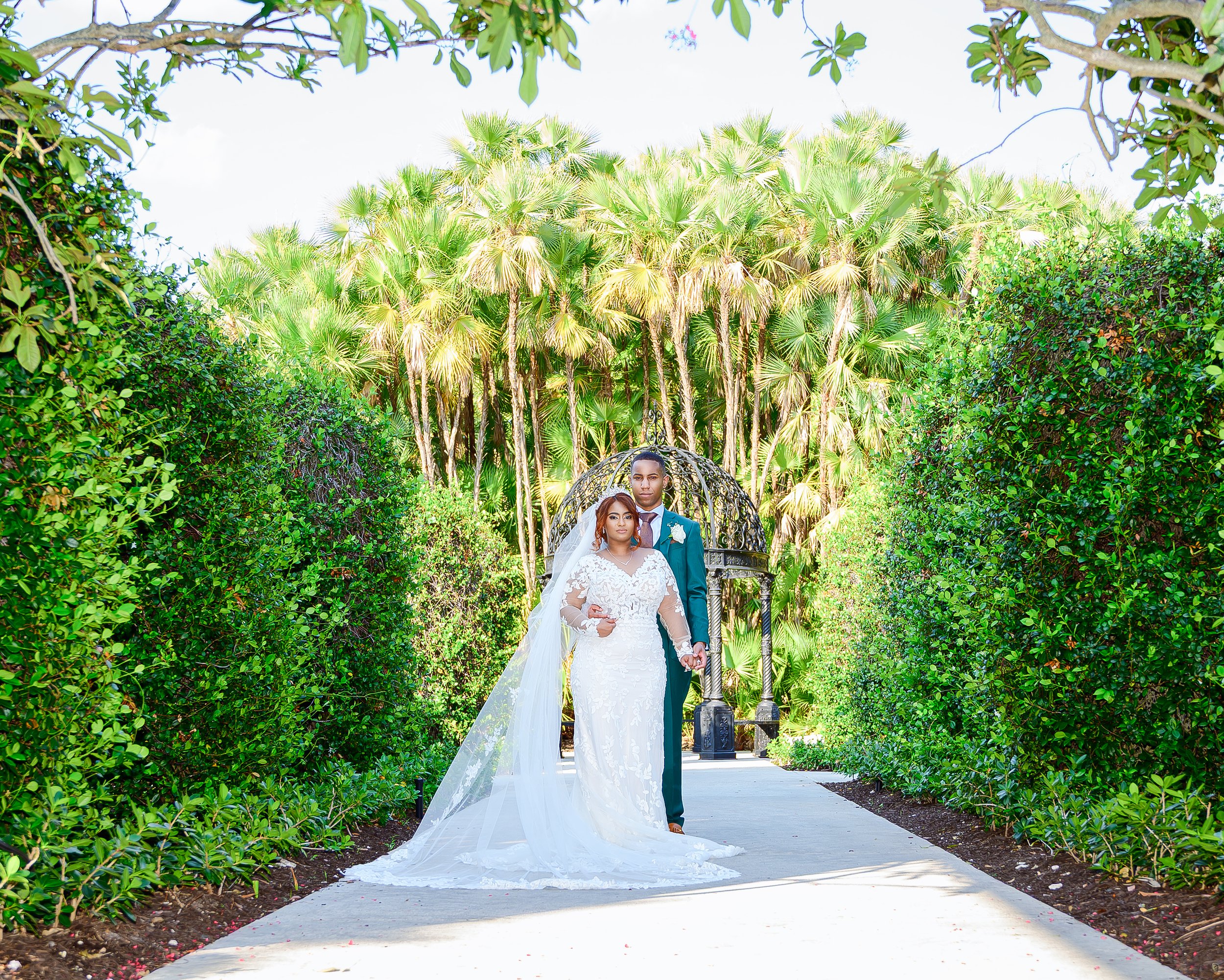A bride and groom standing together on a garden path surrounded by lush green bushes and trees, with palm trees in the background, during a wedding photoshoot.