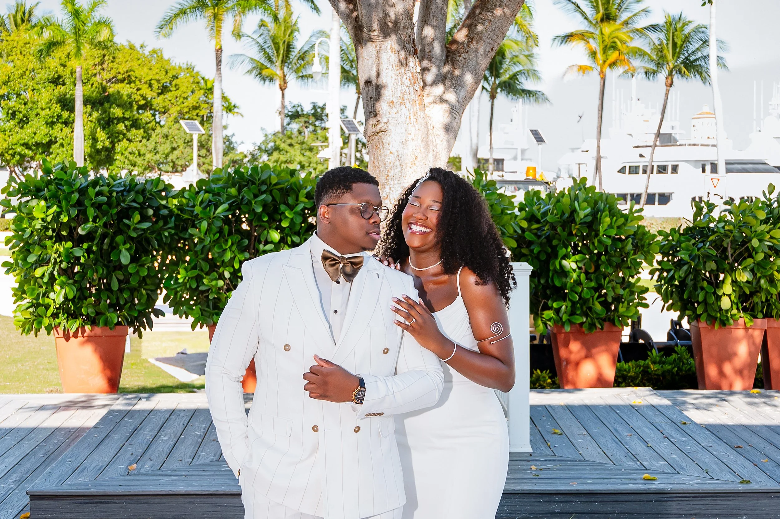 A smiling woman in a white dress is resting her hand on the shoulder of a man in a white pinstripe suit and glasses. Both are standing outdoors near a large tree with boats in the background.