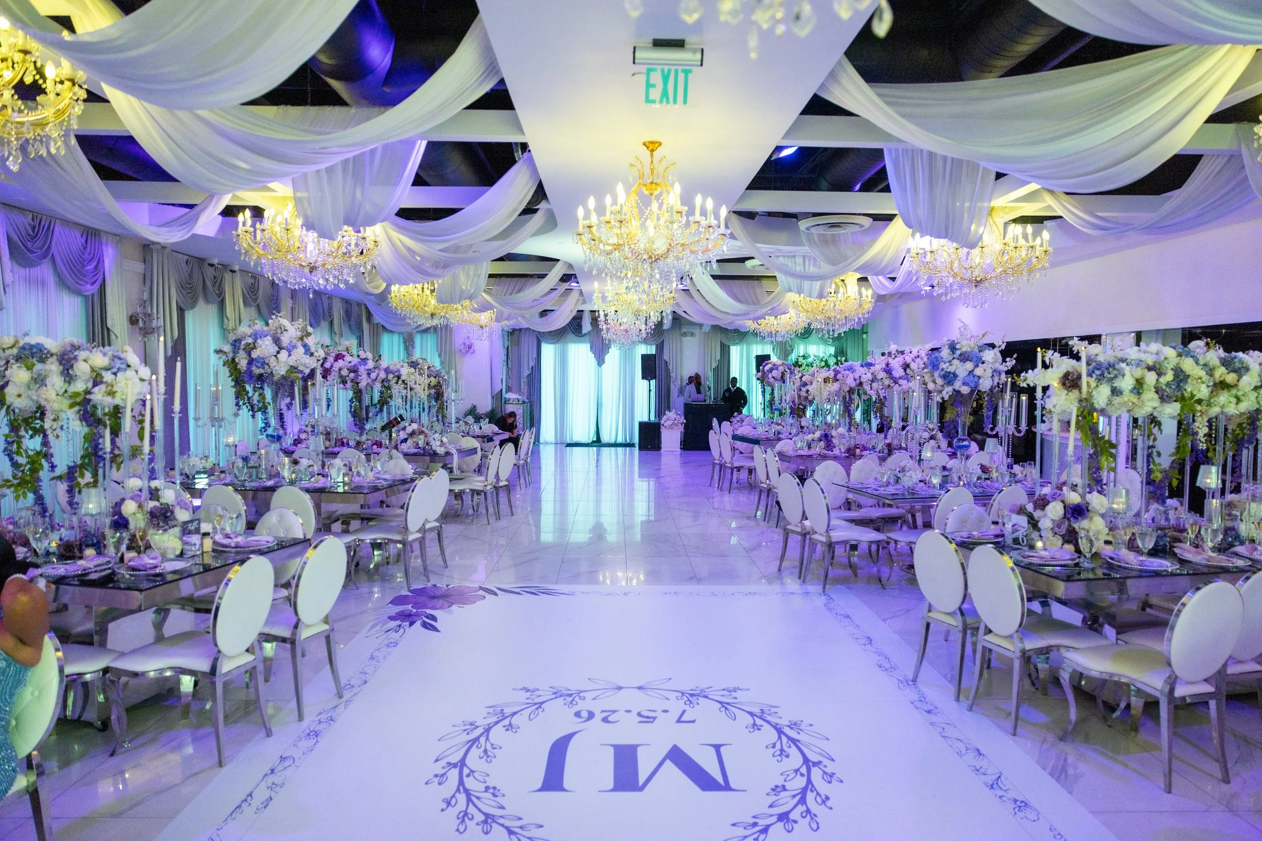 Elegant banquet hall decorated with purple and white flowers, draped white fabric, and multiple chandeliers, set up for a formal event with round tables and white chairs.
