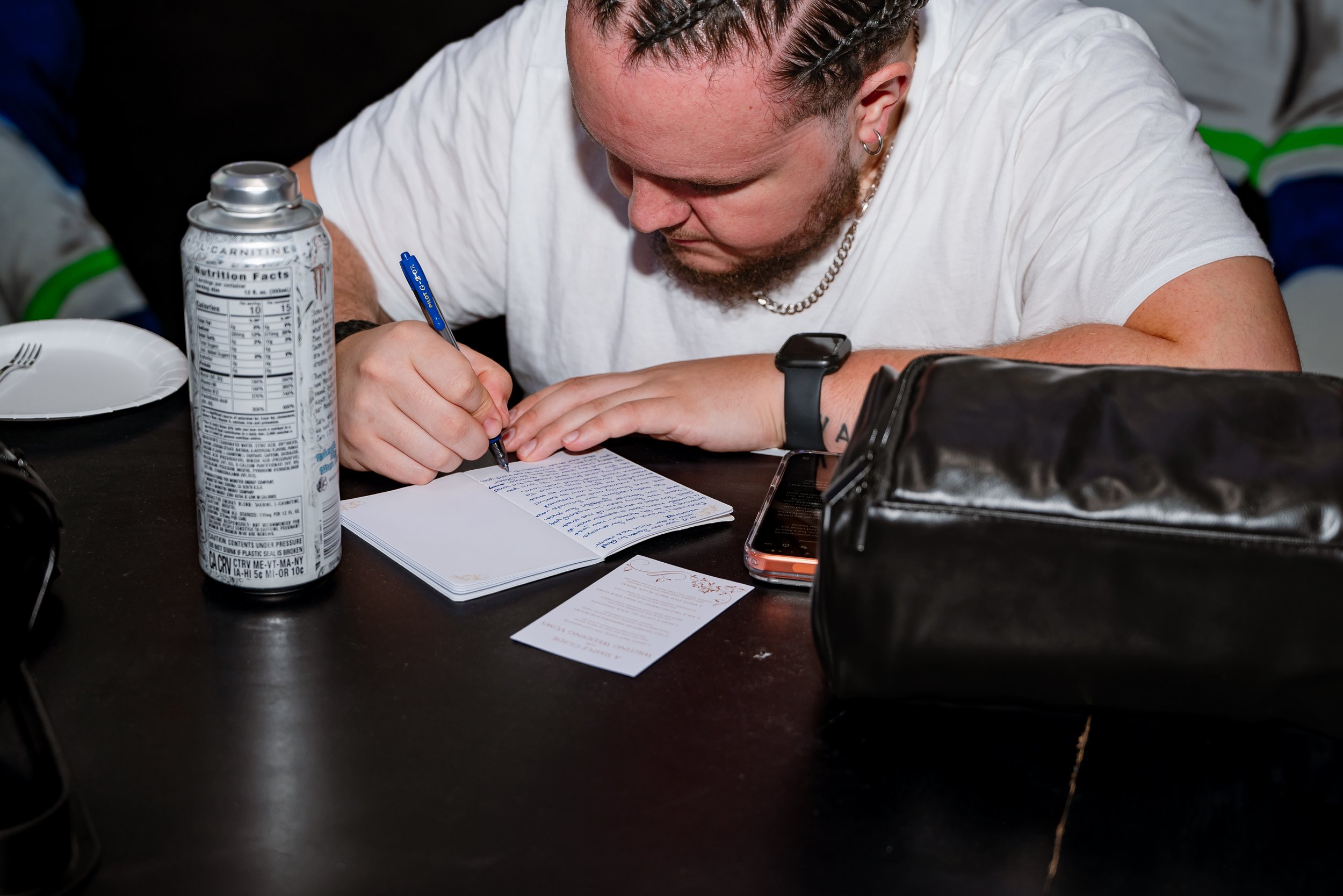 Man with short hair, beard, and earrings writing in a notebook at a desk, surrounded by a soda can, phone, receipt, black bag, and plates.