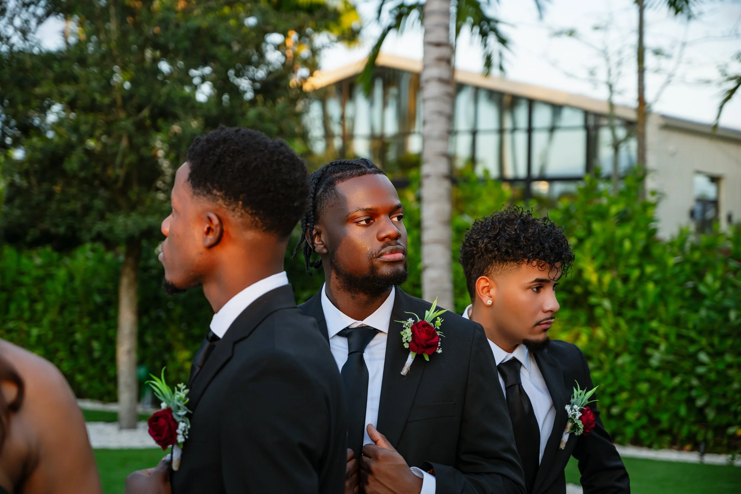 Three young men dressed in black tuxedos with white shirts and black ties, standing outdoors among green foliage and trees, participating in a formal event.