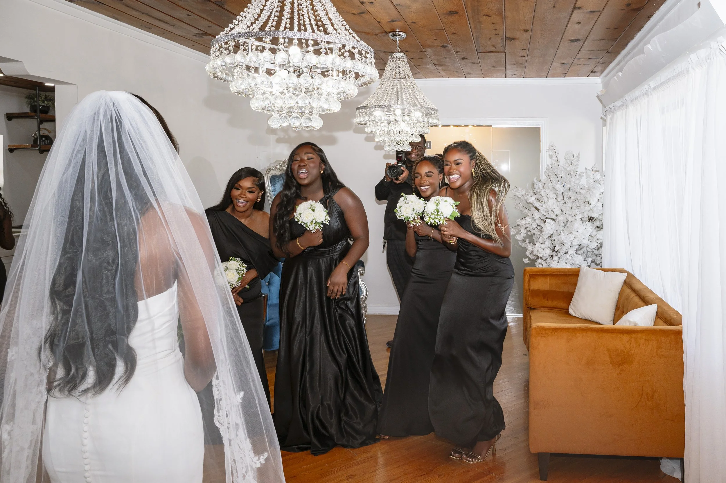 A bride with a long veil and white dress facing four bridesmaids in black dresses, all smiling and holding bouquets of white flowers, inside a decorated room with chandeliers and white curtains.