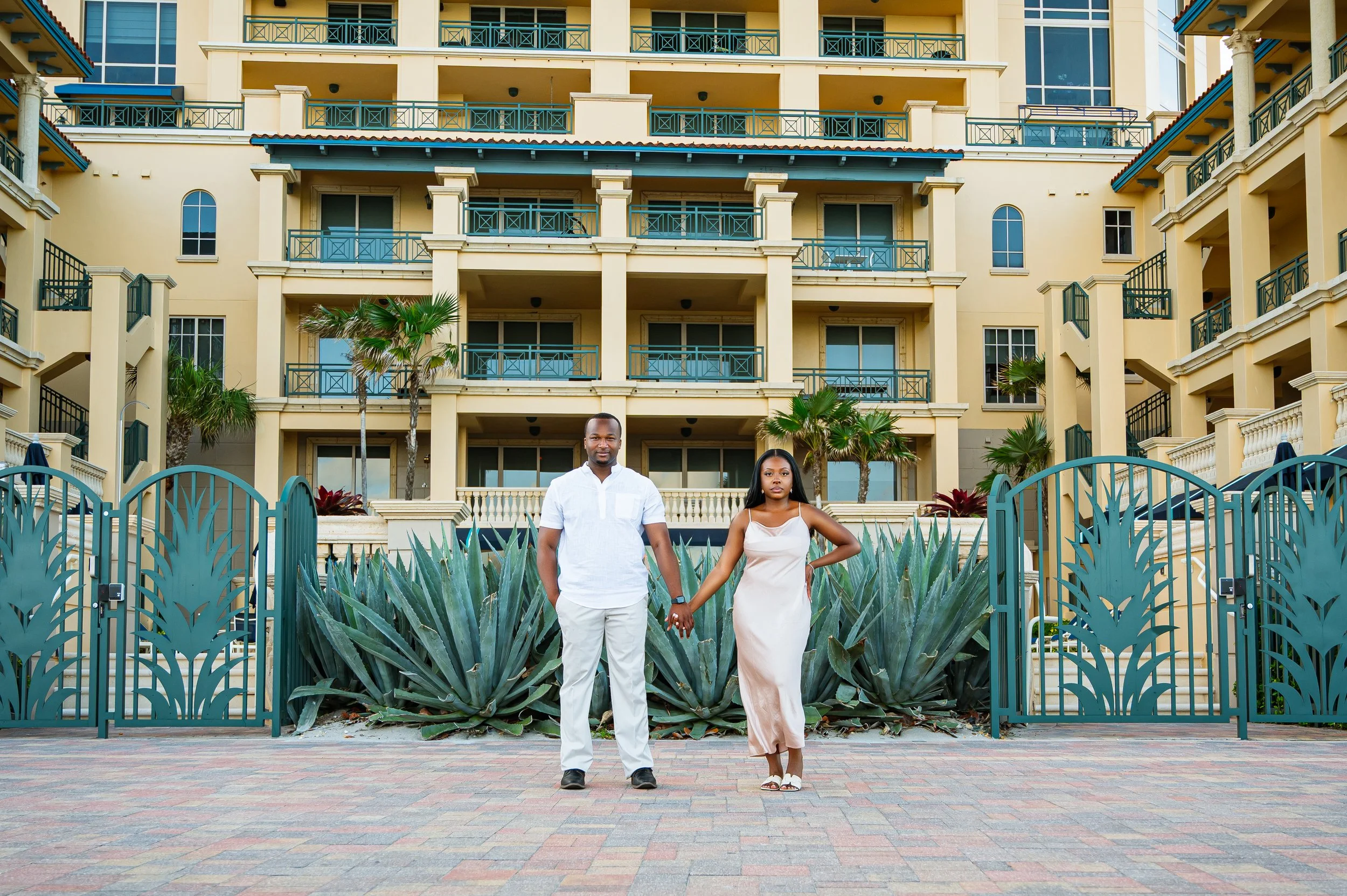 A couple holding hands stands in front of a yellow multi-story building with balconies and palm trees. The man is wearing an all-white outfit, and the woman is wearing a dress.
