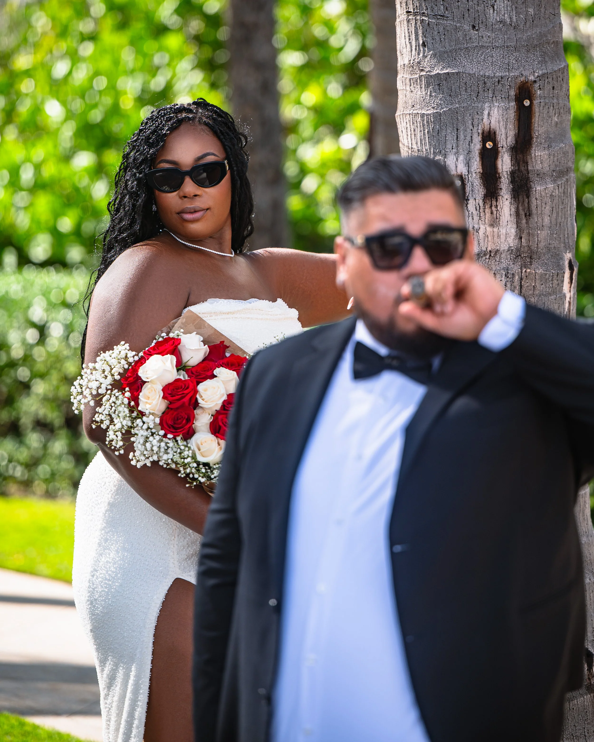 A woman in a white strapless dress holding a bouquet of red and white roses and wearing sunglasses, standing behind a man in a tuxedo and sunglasses, outdoors near a tree.