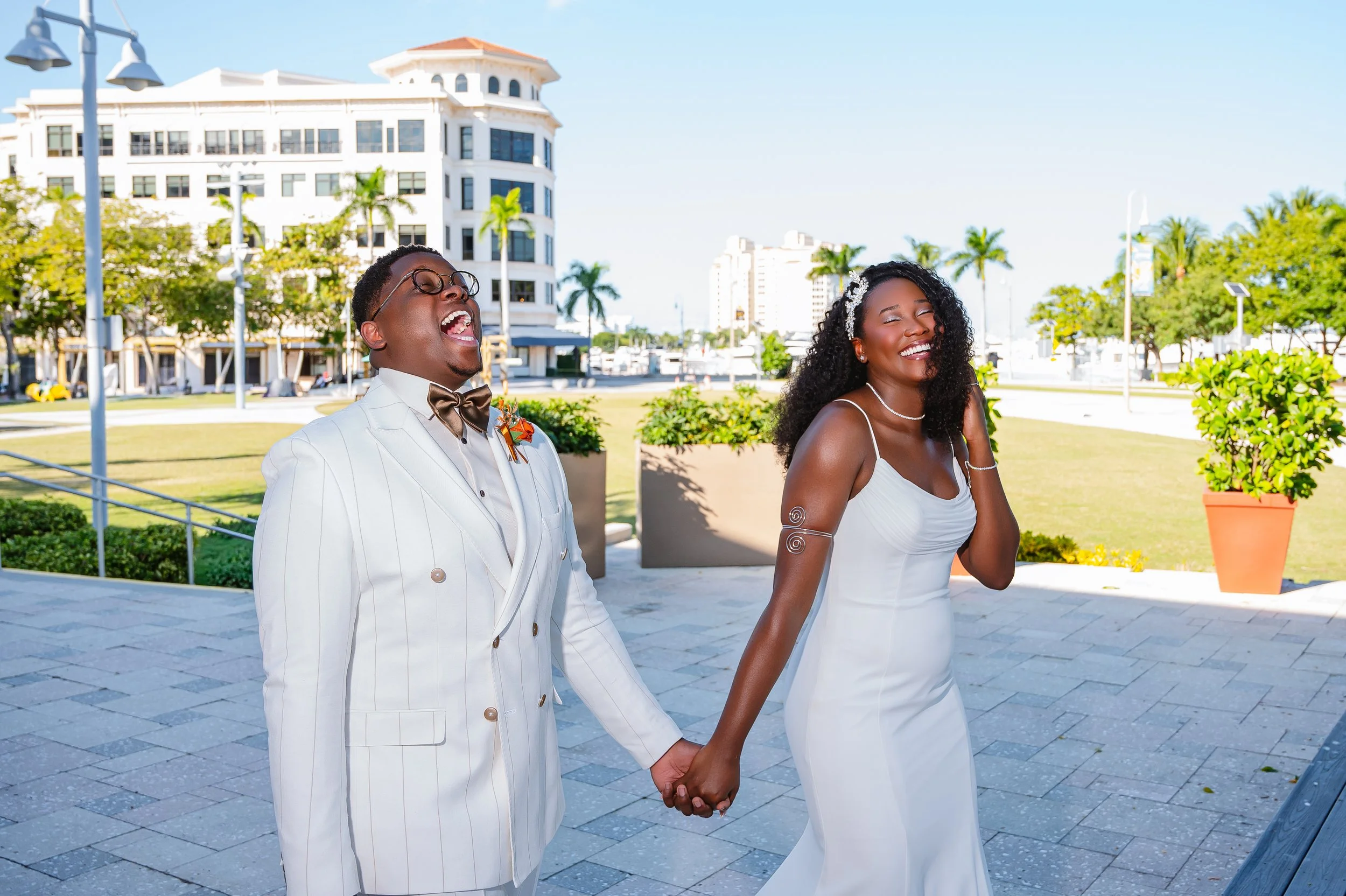 A newlywed couple dressed in white wedding attire holding hands and laughing outdoors on a sunny day with modern buildings and palm trees in the background.