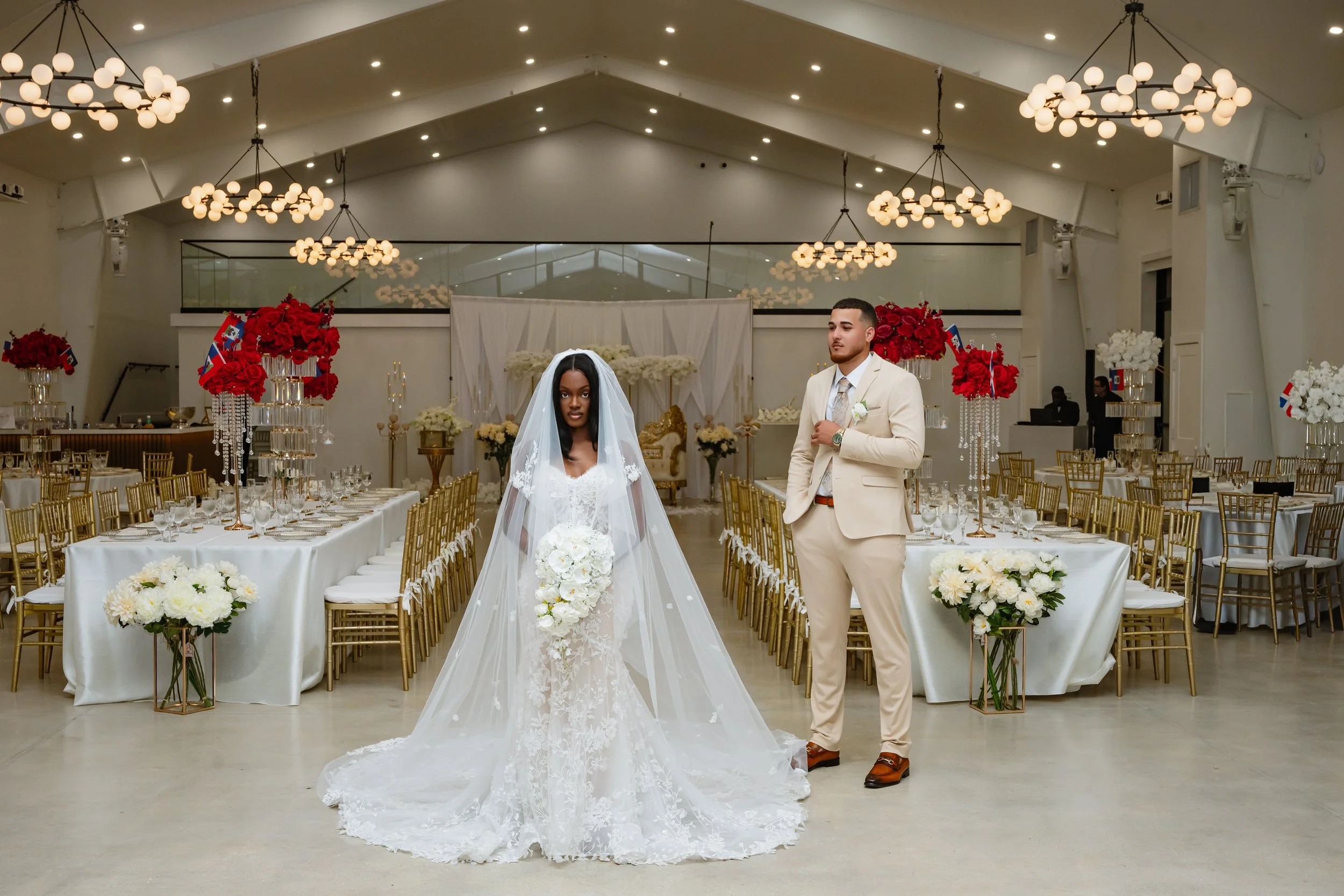 A bride in a white wedding gown and veil stands beside a groom in a beige suit inside a decorated wedding reception hall with long tables and floral arrangements.