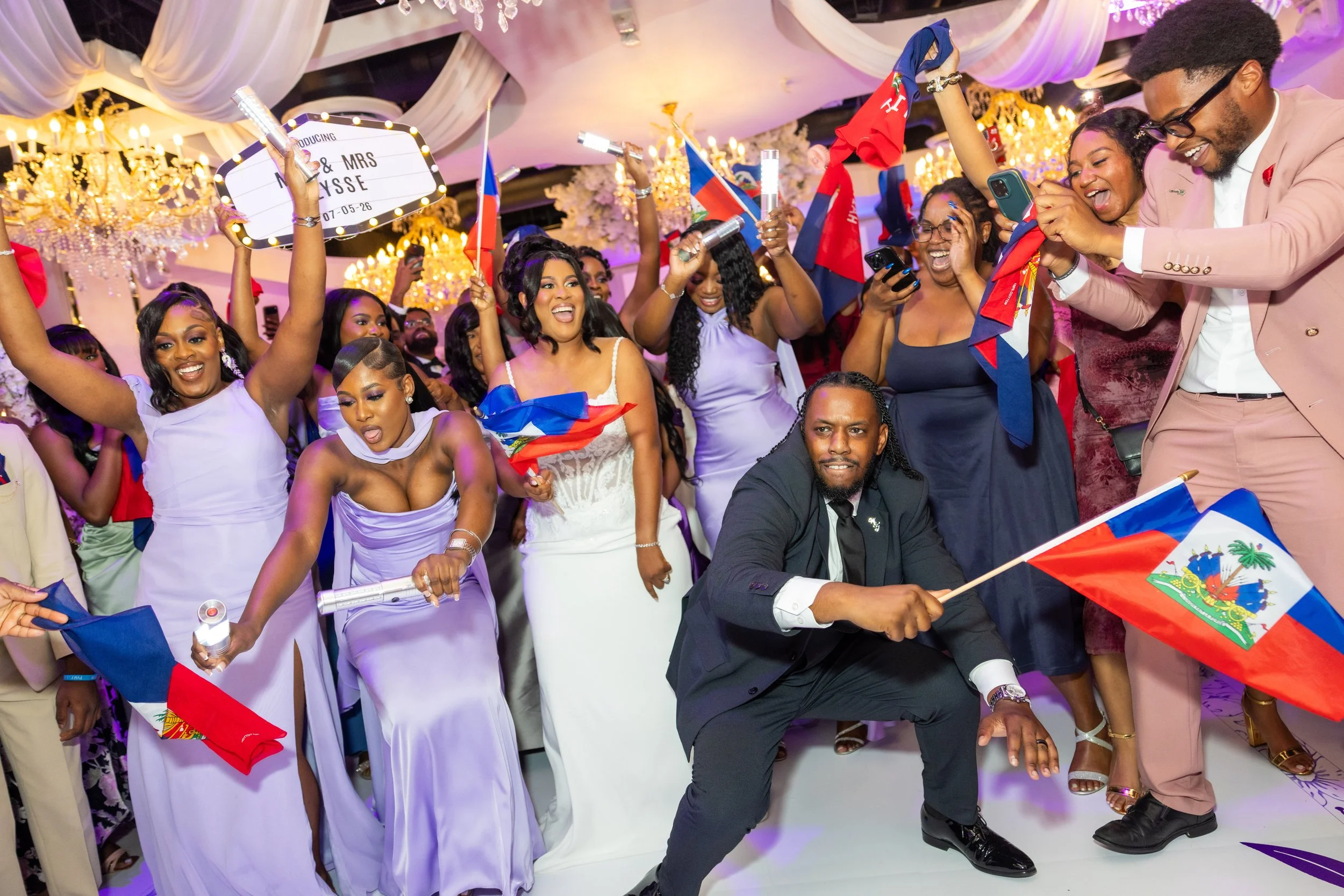 A group of people celebrating at an event with Haitian flags, smiling, cheering, and taking photos in a decorated banquet hall with chandeliers and purple lighting.