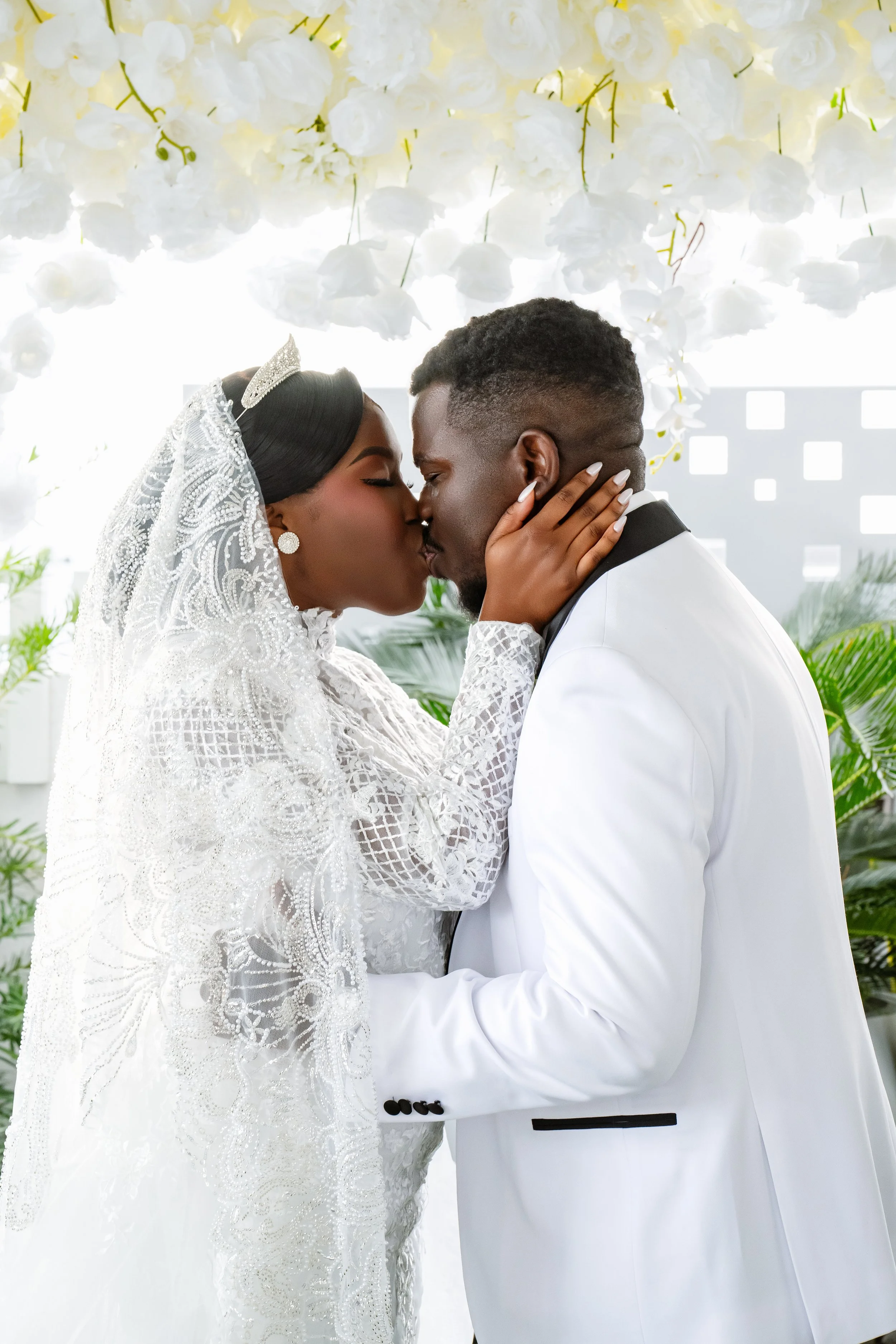 A bride and groom kissing during their wedding ceremony, with the bride wearing a lace wedding dress and a tiara, and the groom in a white suit, under a floral canopy of white flowers.