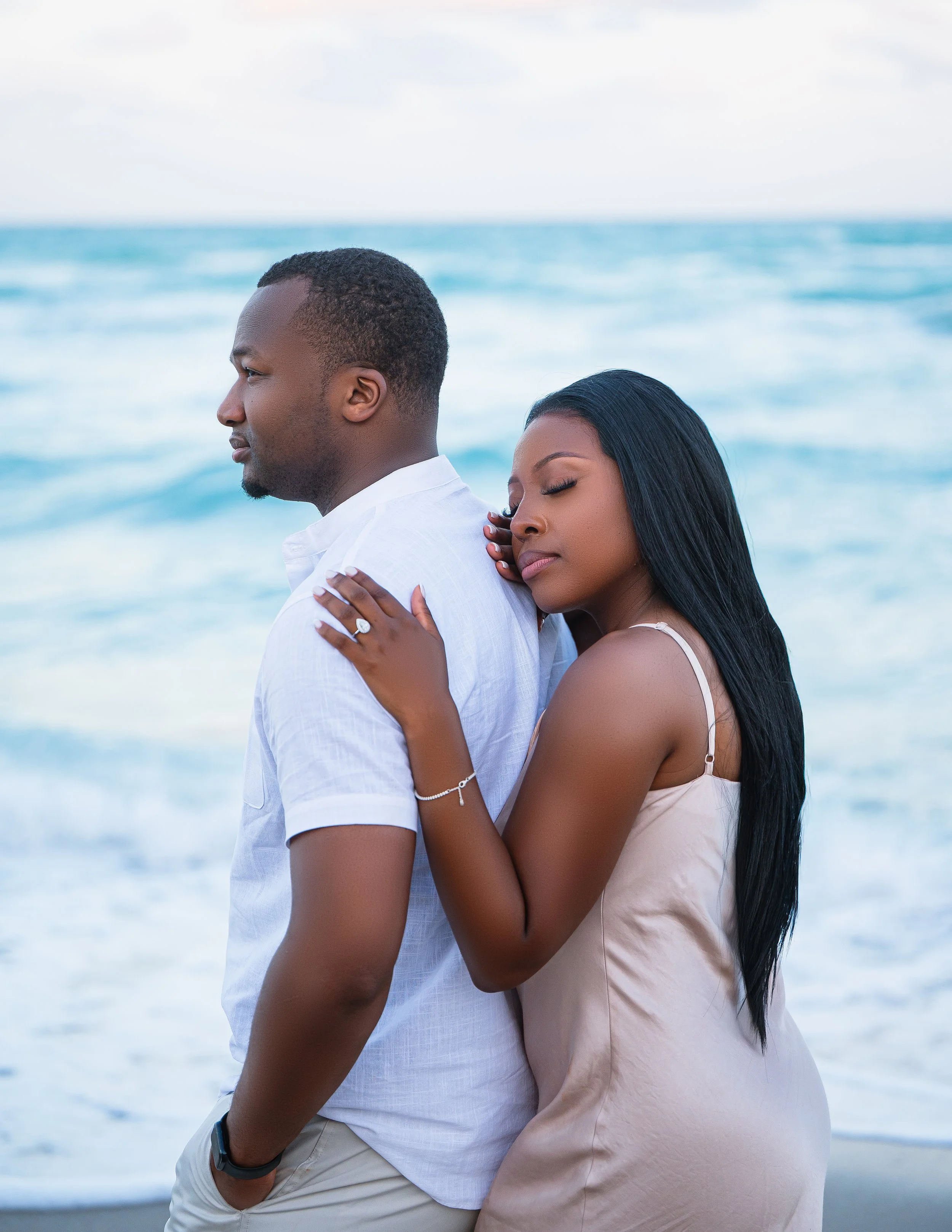 A couple stands on the beach by the ocean. The woman is hugging the man from behind with her eyes closed, and she is wearing a beige dress and jewelry. The man is wearing a white shirt with his hands in his pockets.