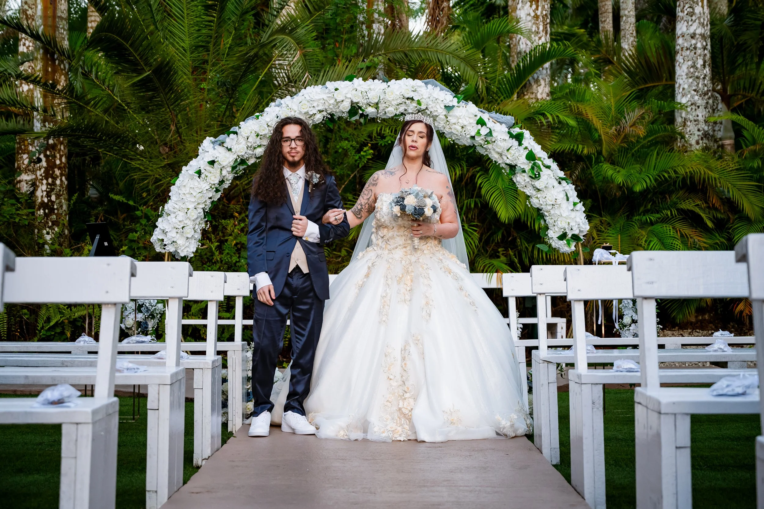 A bride and groom stand under a white floral arch at an outdoor wedding ceremony. The bride wears a white ball gown with gold embroidery and a veil, holding a bouquet of white and blue flowers. The groom wears a dark blue tuxedo with a beige vest and