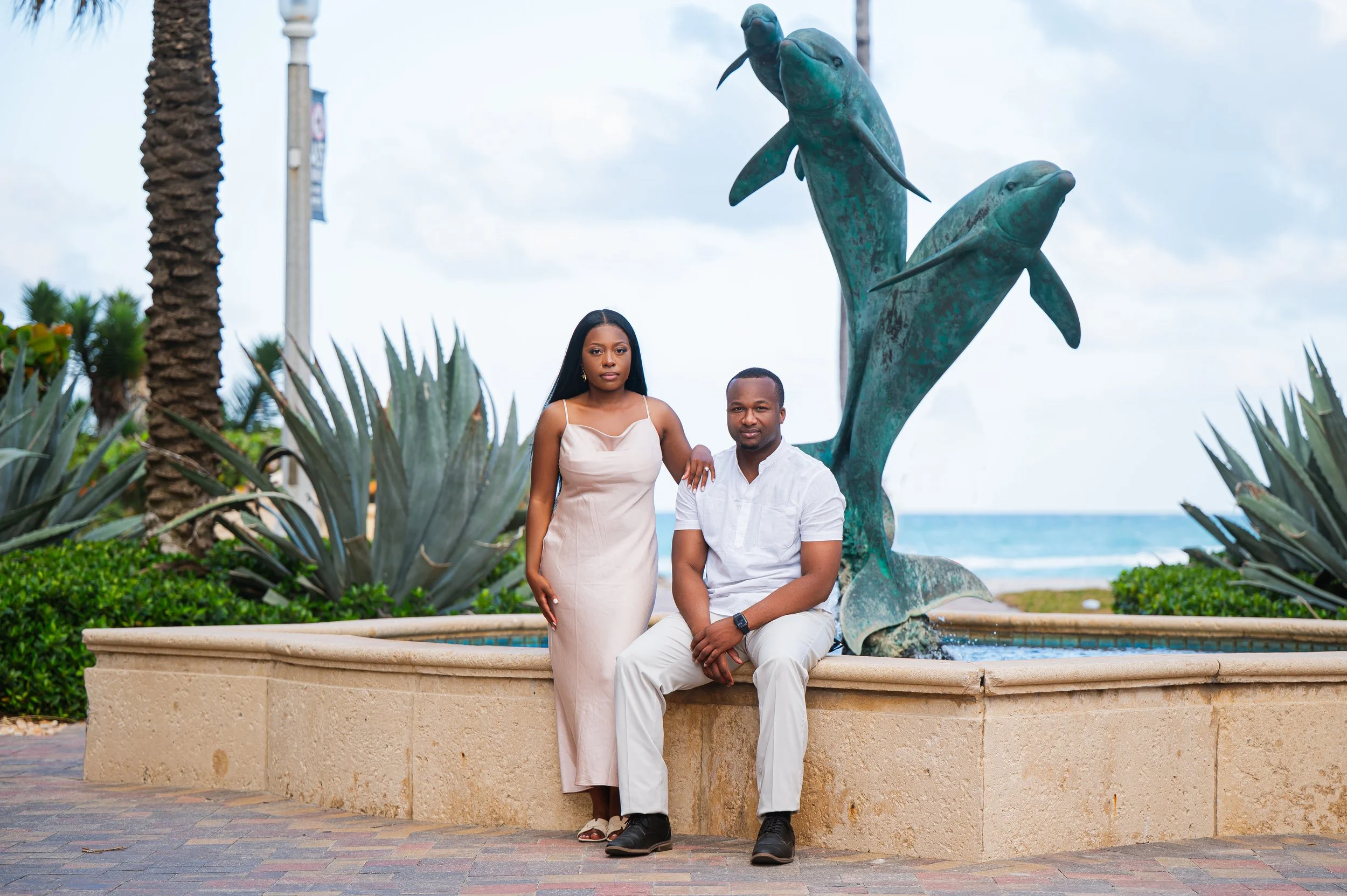 A woman and a man in front of a dolphin fountain on a beachside promenade, with the ocean in the background.