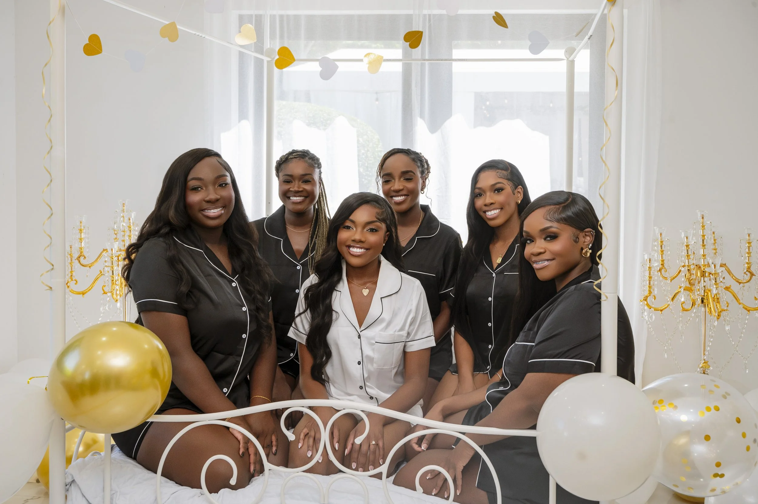 Group of six women in pajamas sitting on a bed decorated with gold and white balloons and heart-shaped paper garland, smiling at the camera during a celebration in a bright room.
