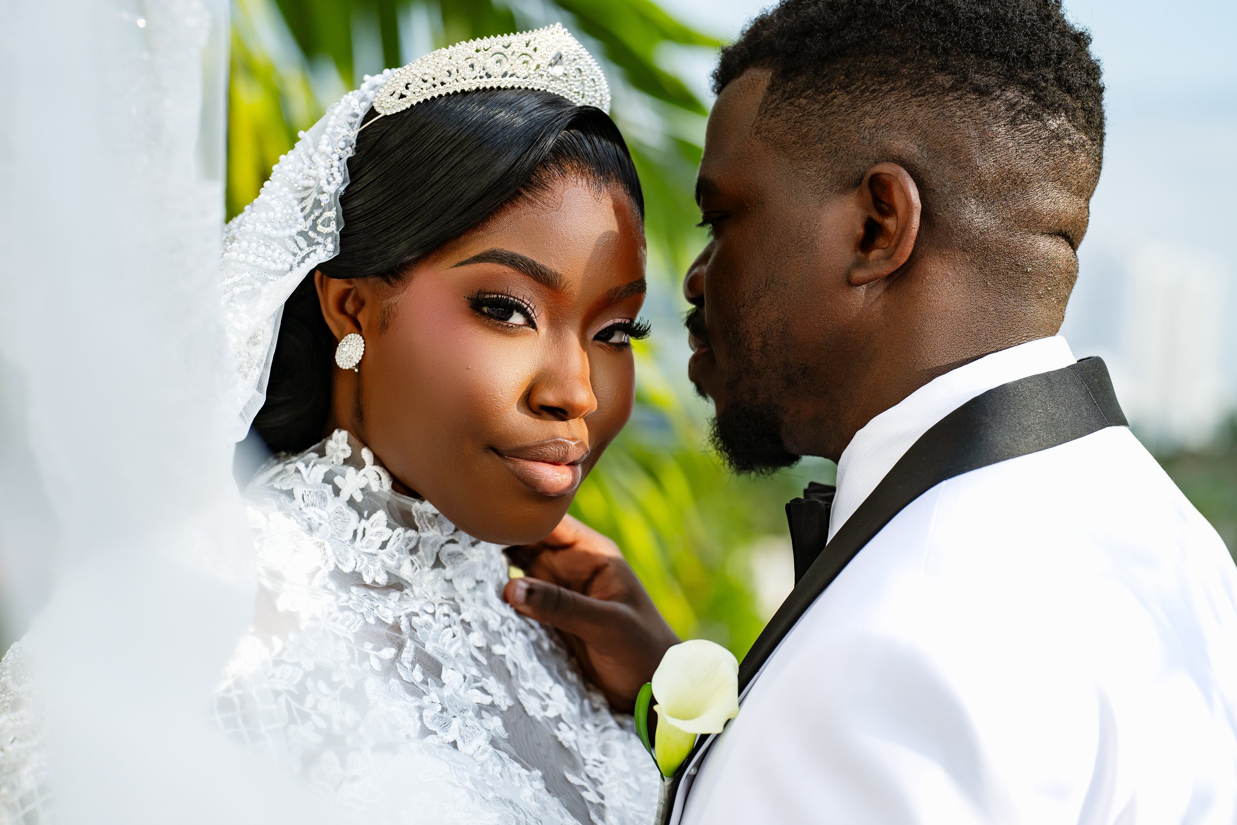 A bride and groom close together outdoors, with the bride wearing a white lace dress and a decorative tiara, and the groom in a black tuxedo with a white shirt and black bow tie, surrounded by green foliage.