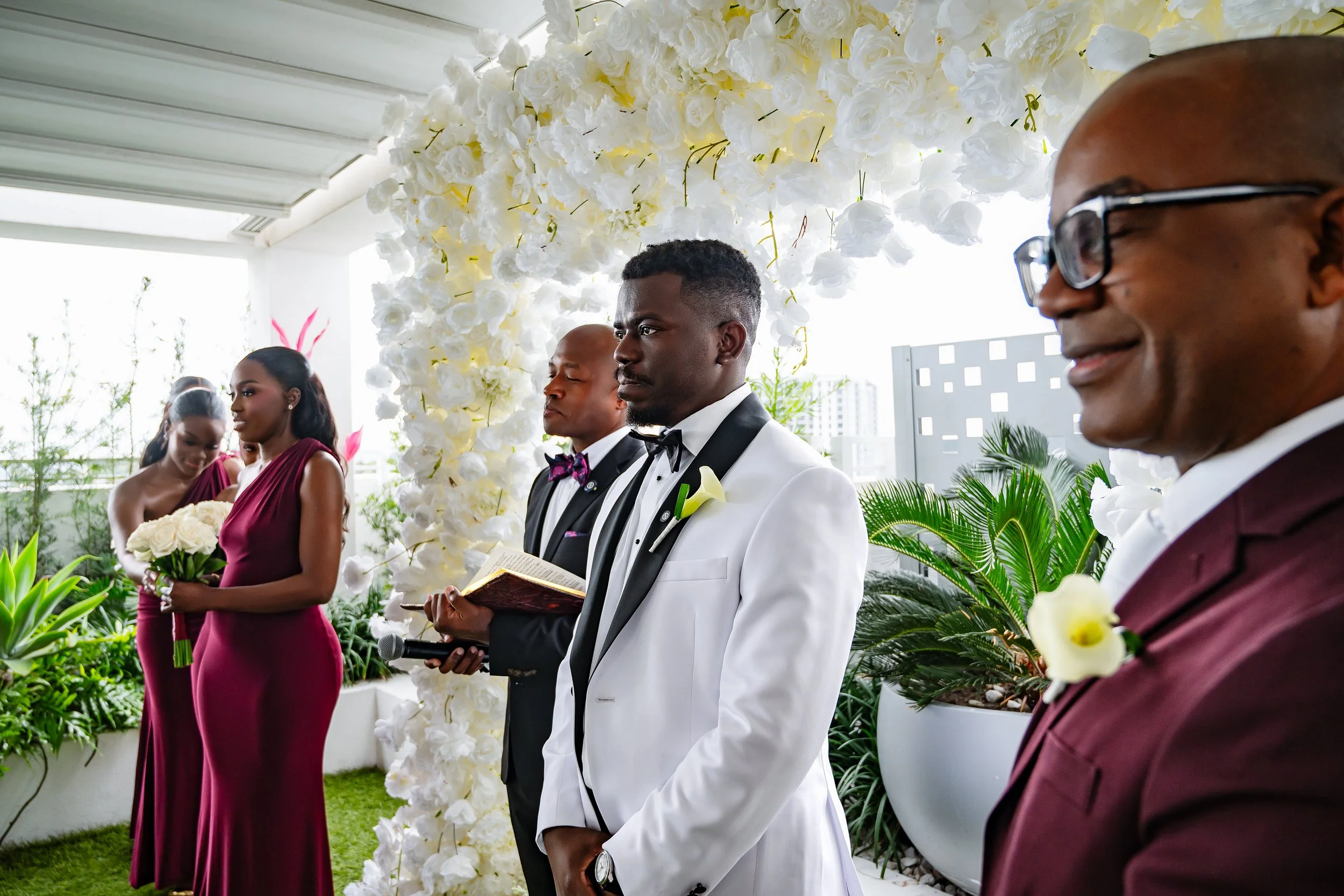 A wedding ceremony with four people standing in front of a floral arch on a balcony, with city buildings in the background. The groom is in a white tuxedo with a black bowtie, holding a book. The woman behind him is in a burgundy dress, and two other